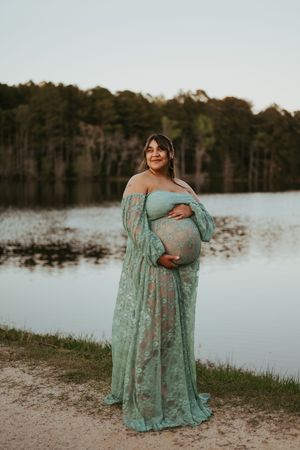 Pregnant woman in teal lace maternity gown cradling baby bump by a serene lake at dusk.