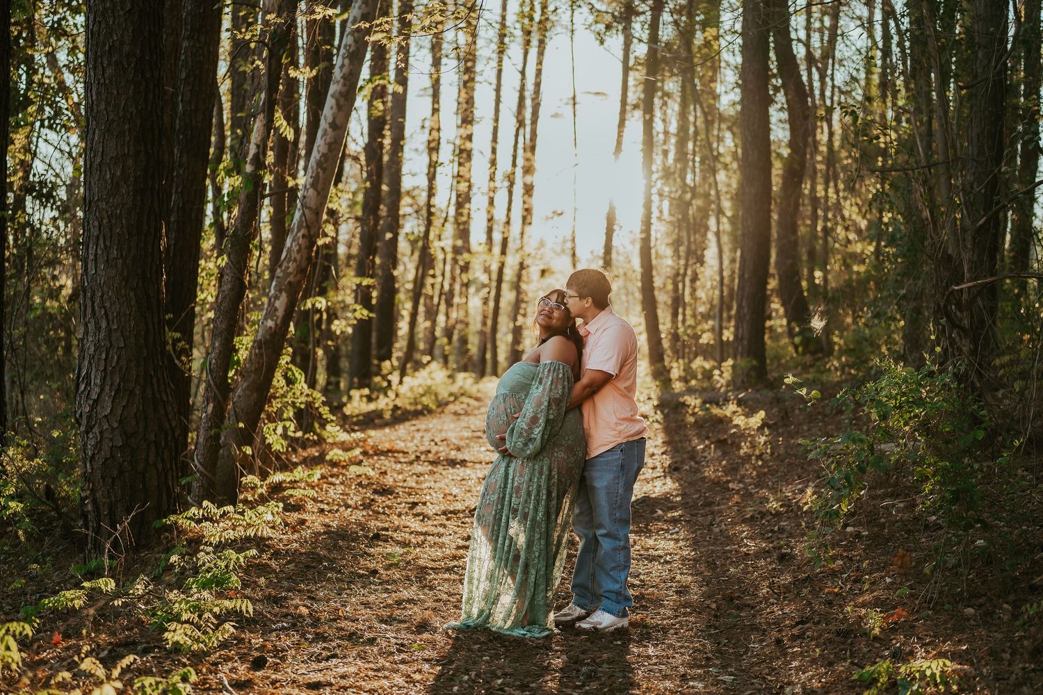 Couple embracing on a sunlit forest path, woman in flowing green dress, golden hour light filtering through tall trees.