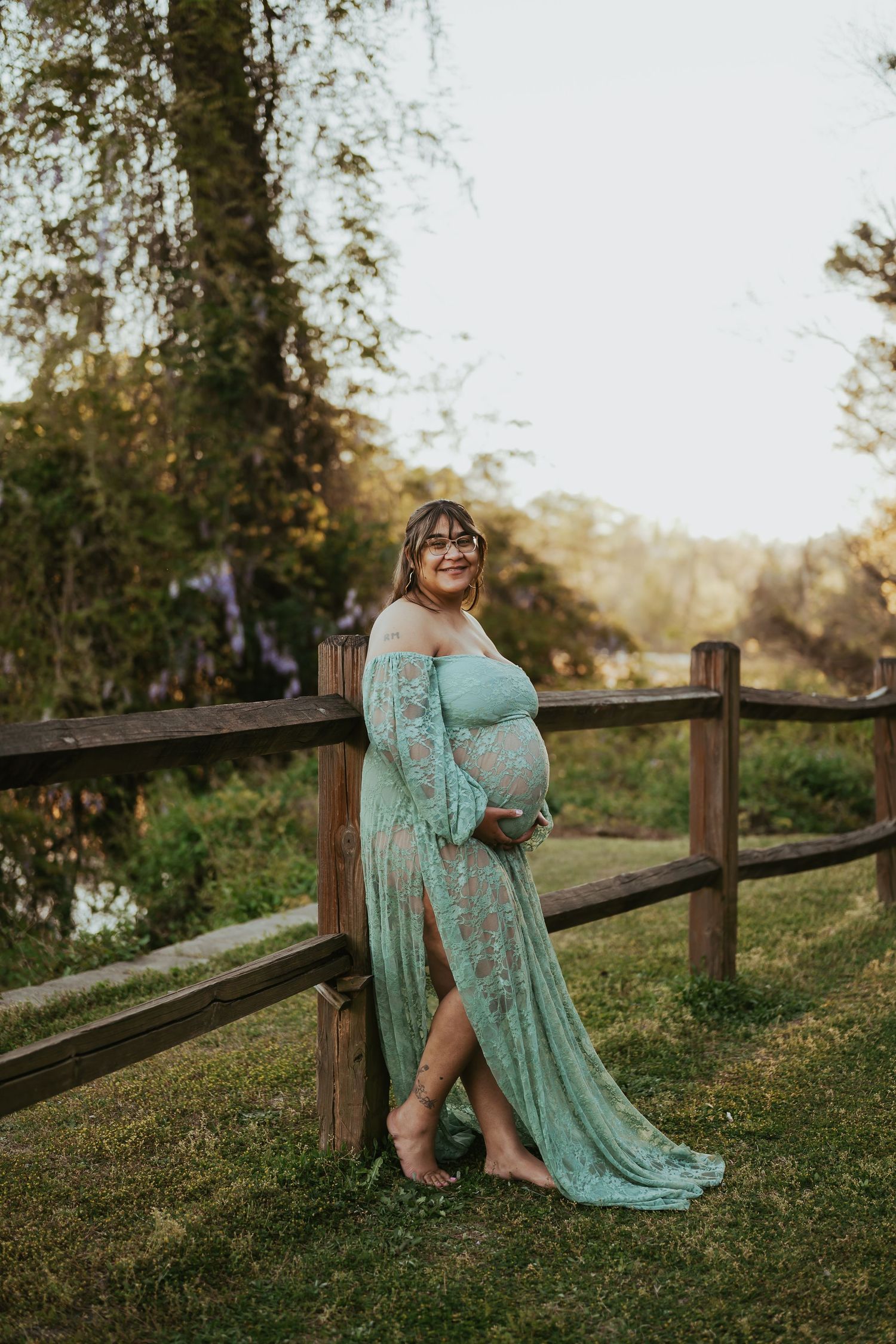 Pregnant woman in teal lace maternity gown leaning on wooden fence outdoors at golden hour.
