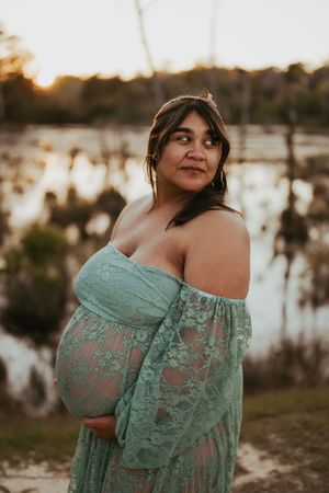 Pregnant woman in teal lace off-shoulder gown poses by a serene lake at golden hour sunset.