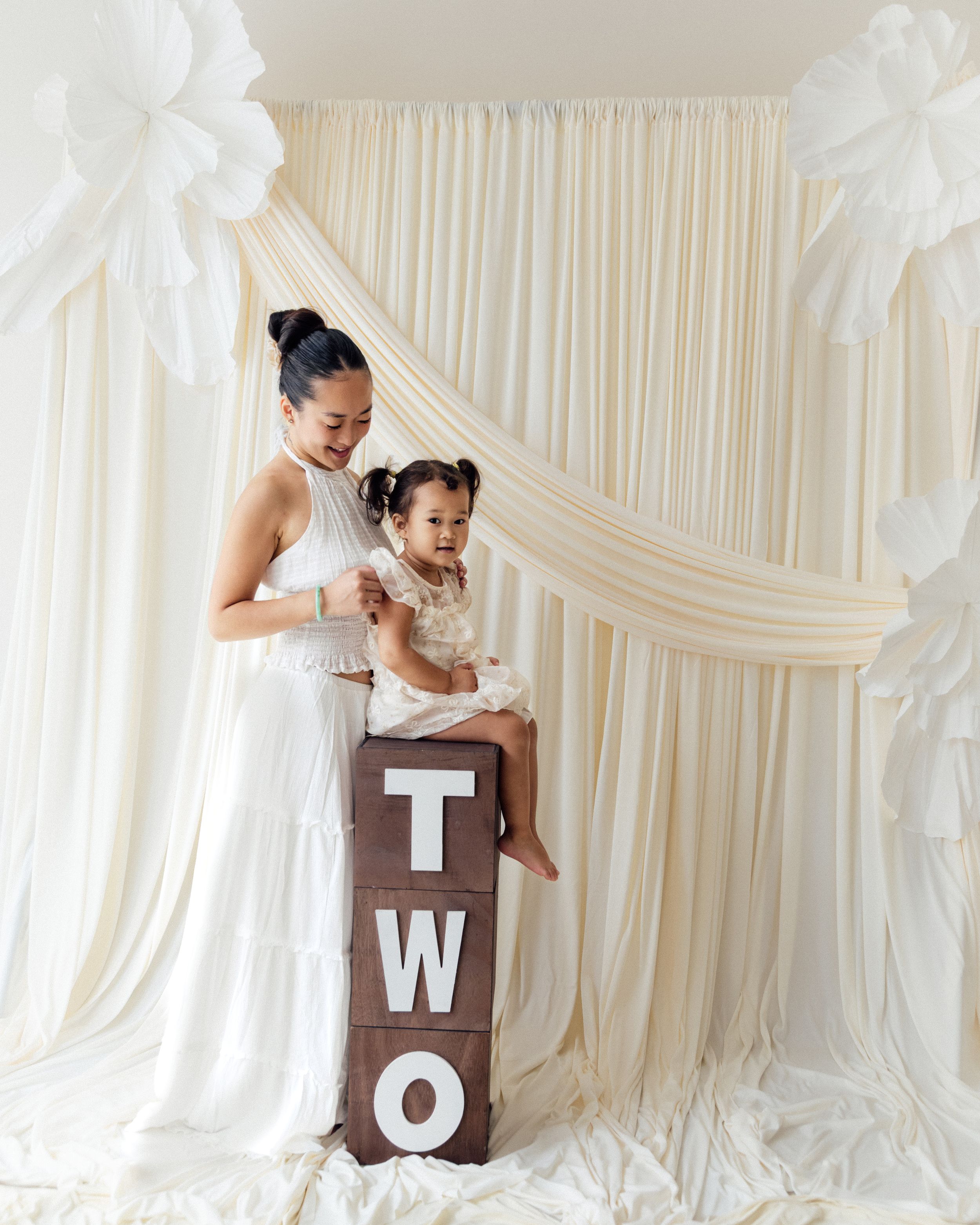 Mother in white dress holds toddler on wooden TWO blocks at elegant cream draped birthday backdrop.