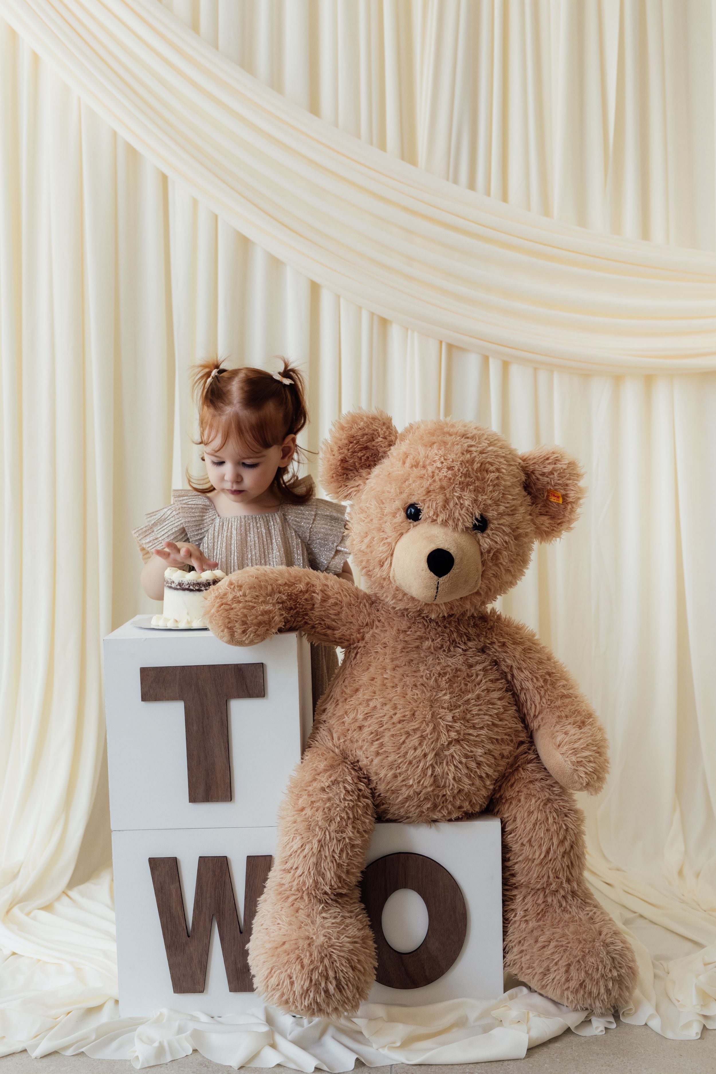 Toddler girl at second birthday party with large teddy bear and TWO letter blocks with small cake.