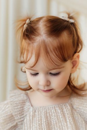 A young girl with red hair in pigtails wearing a sparkly dress looks down sweetly.