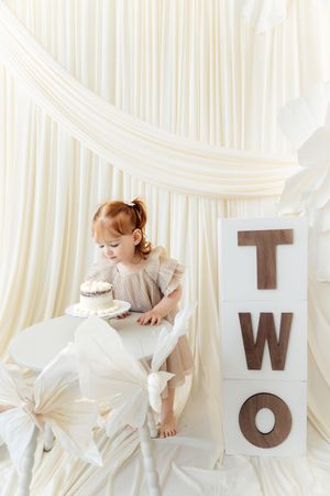 Toddler girl celebrating her 2nd birthday with a cake beside a wooden TWO sign in a white decorated setting.