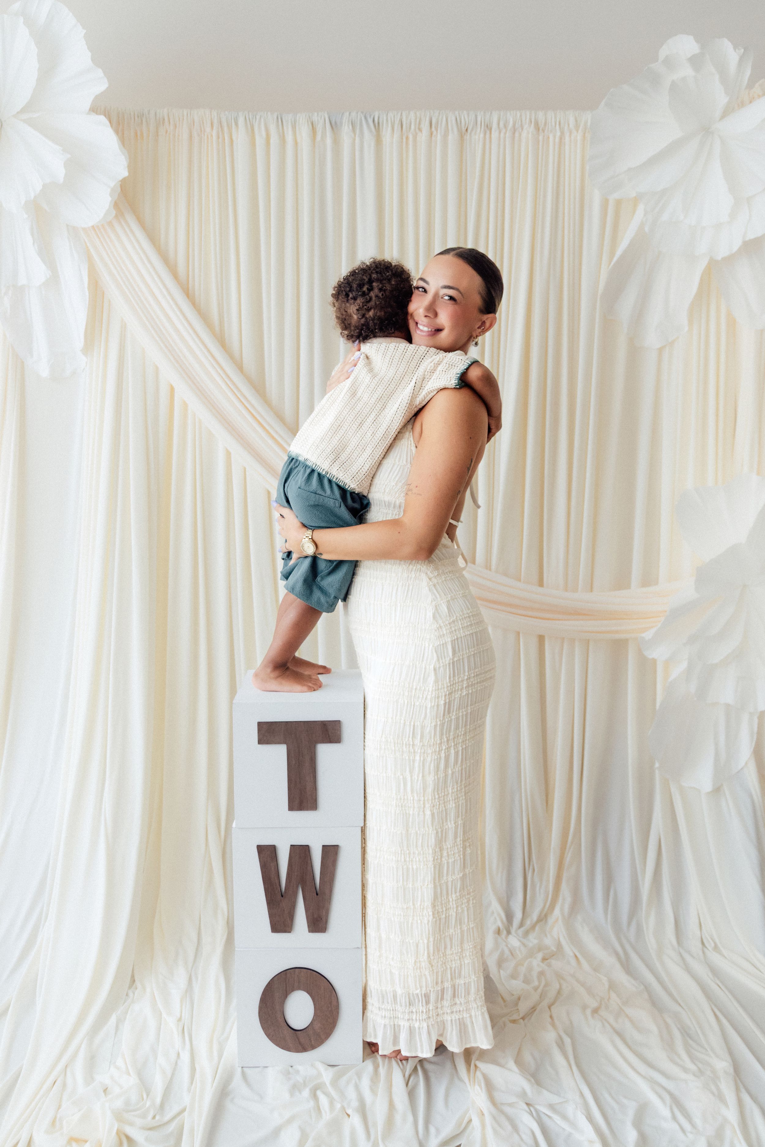 A mother in a white dress holds her toddler standing on TWO letter blocks, posing before a draped cream backdrop.