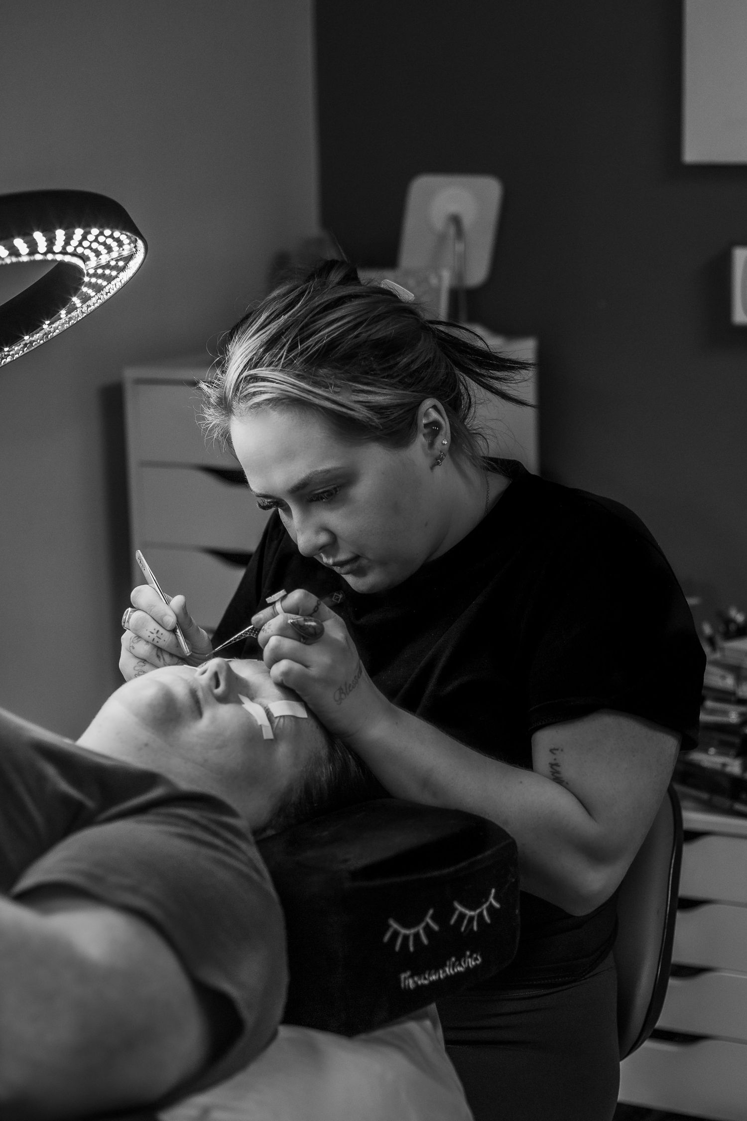 Lash technician applying eyelash extensions to a client in a professional salon, black and white photo.