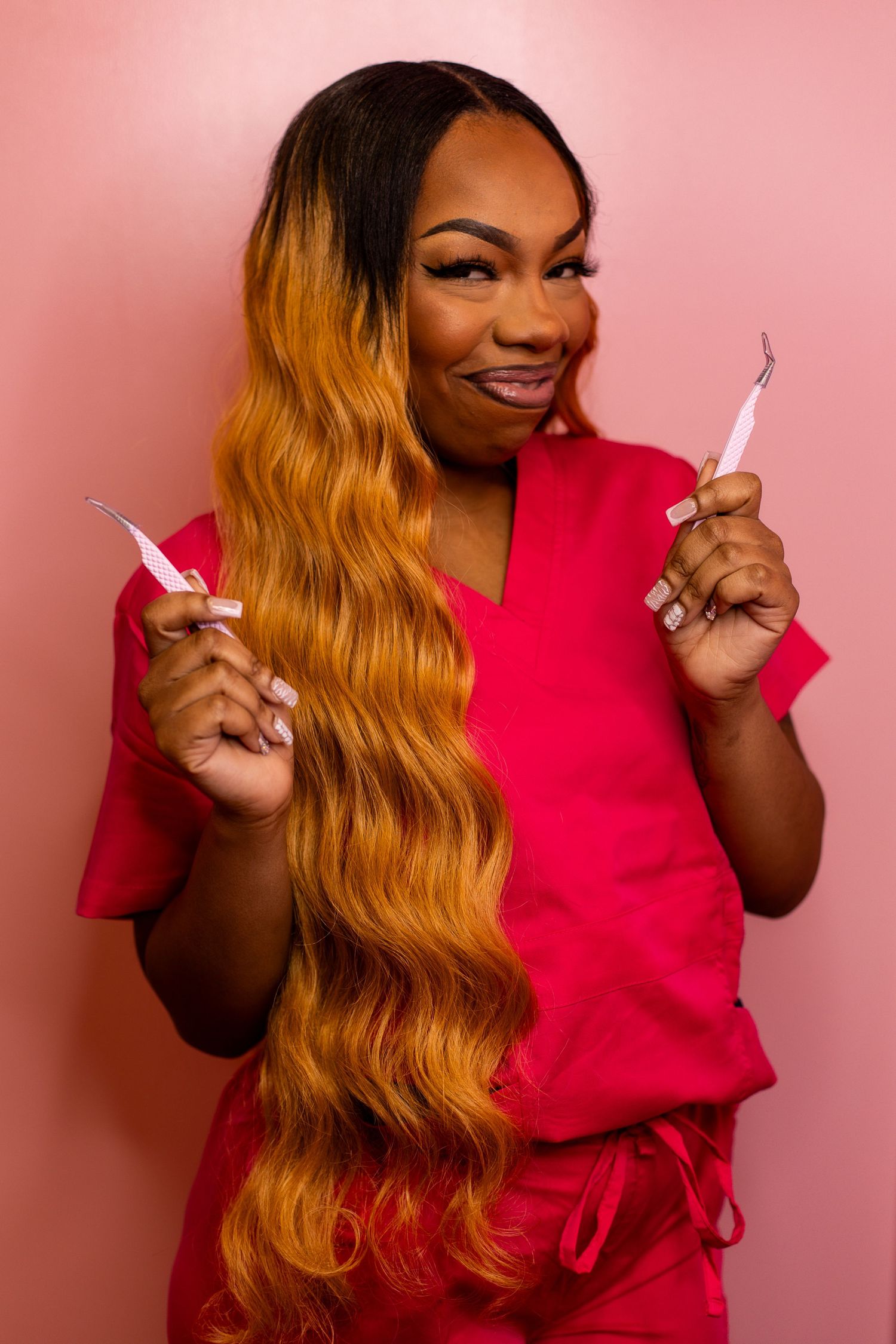 A smiling woman in red scrubs with long ombre hair holds two dental tools against a pink background.