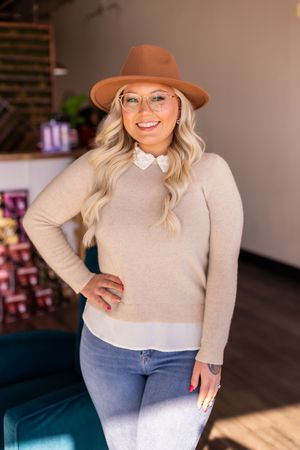 Smiling blonde woman in tan sweater, brown fedora, and glasses posing in a salon.
