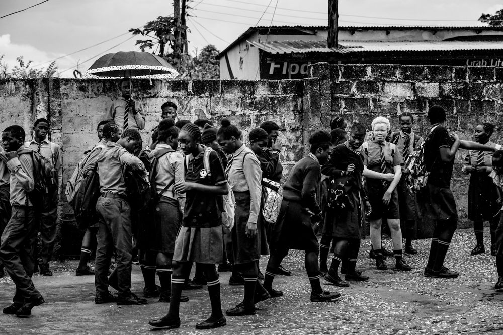 Black and white photo of African schoolchildren in uniforms walking together on a dirt road near a concrete wall.