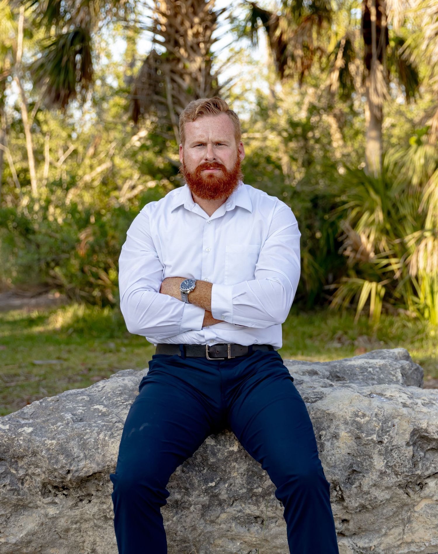 Red-bearded man in white shirt and navy trousers holds jacket over shoulder on a tropical dirt path.