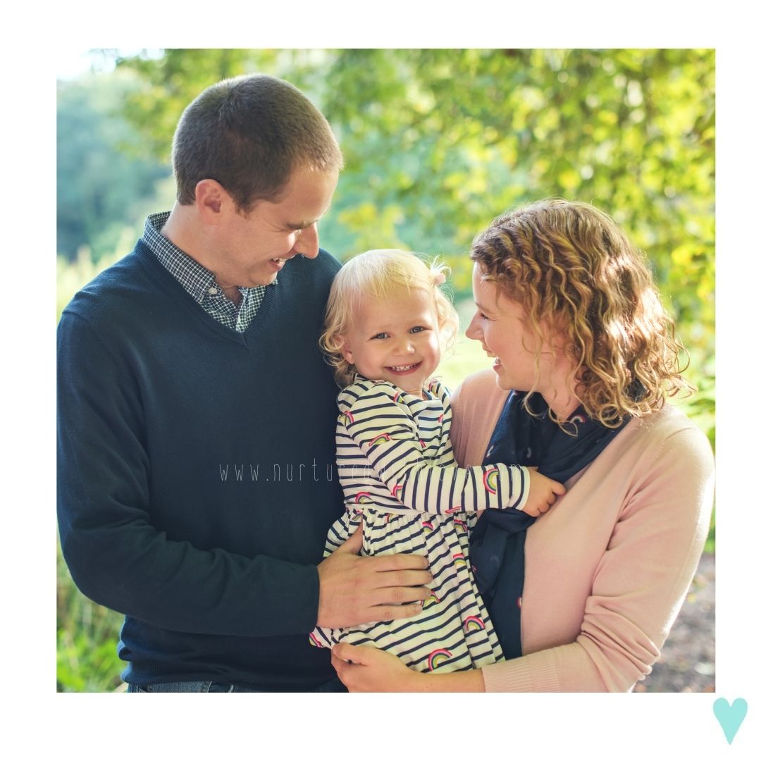 A happy young family with a toddler smiling outdoors, surrounded by lush green trees in warm sunlight.