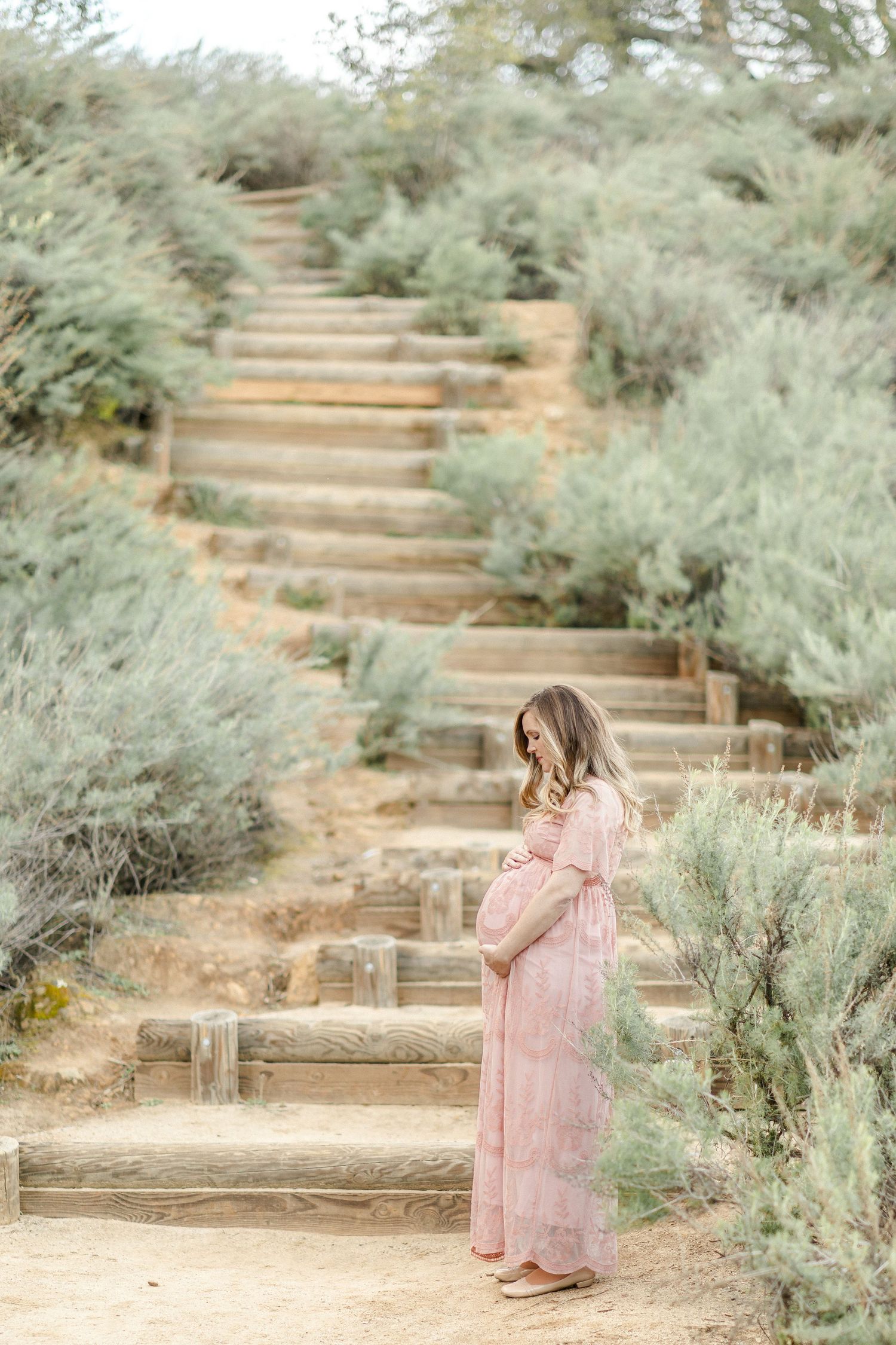 Pregnant woman in pink maxi dress stands on garden steps surrounded by lush green shrubs during outdoor maternity photoshoot.