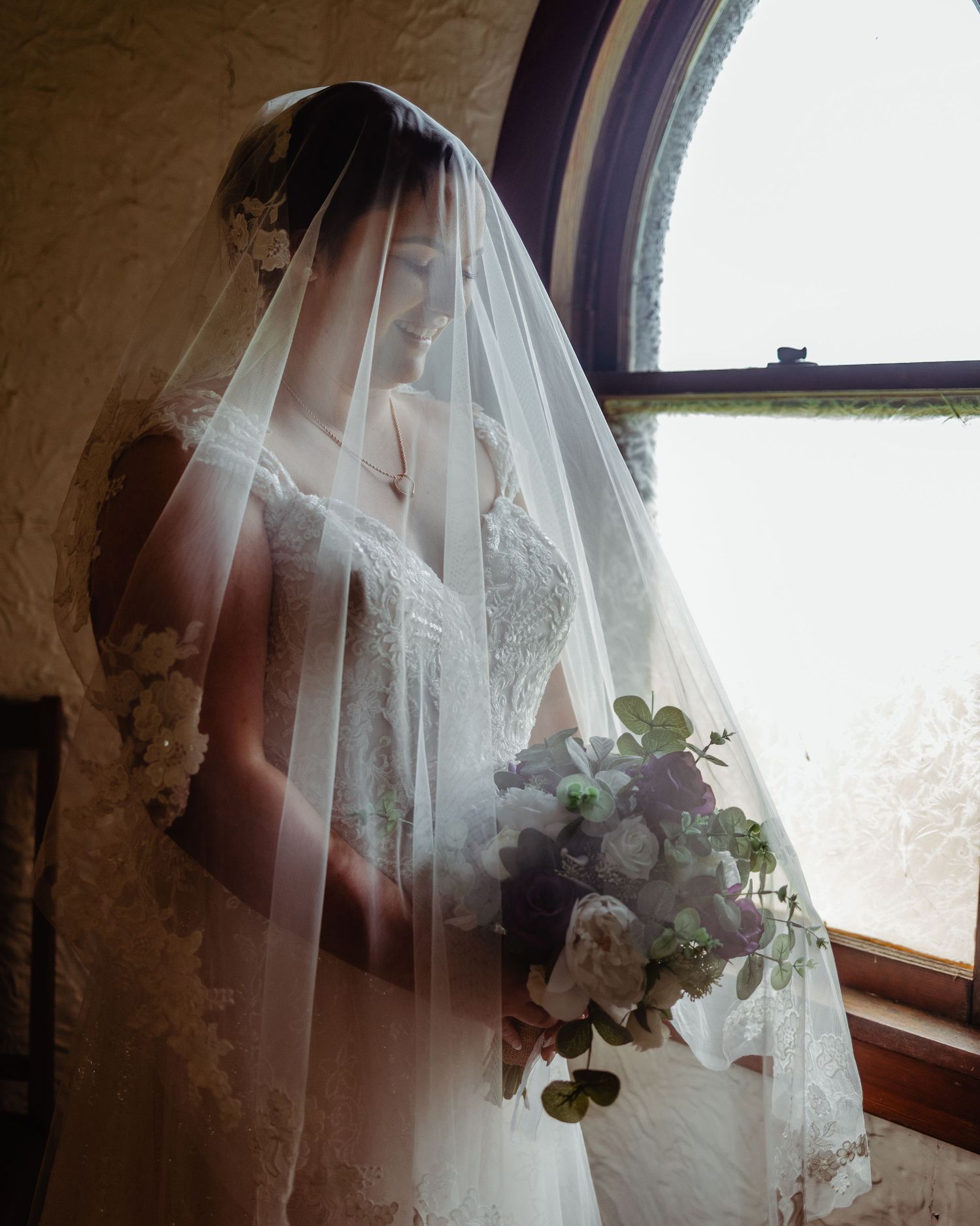 Bride in lace wedding gown and flowing veil holds bouquet of white and purple flowers by arched window.