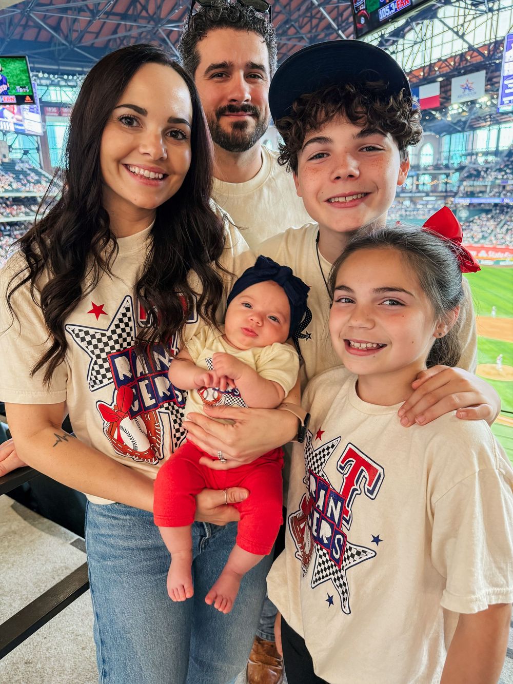 Family of five in matching Texas Rangers shirts posing with newborn baby at baseball stadium.