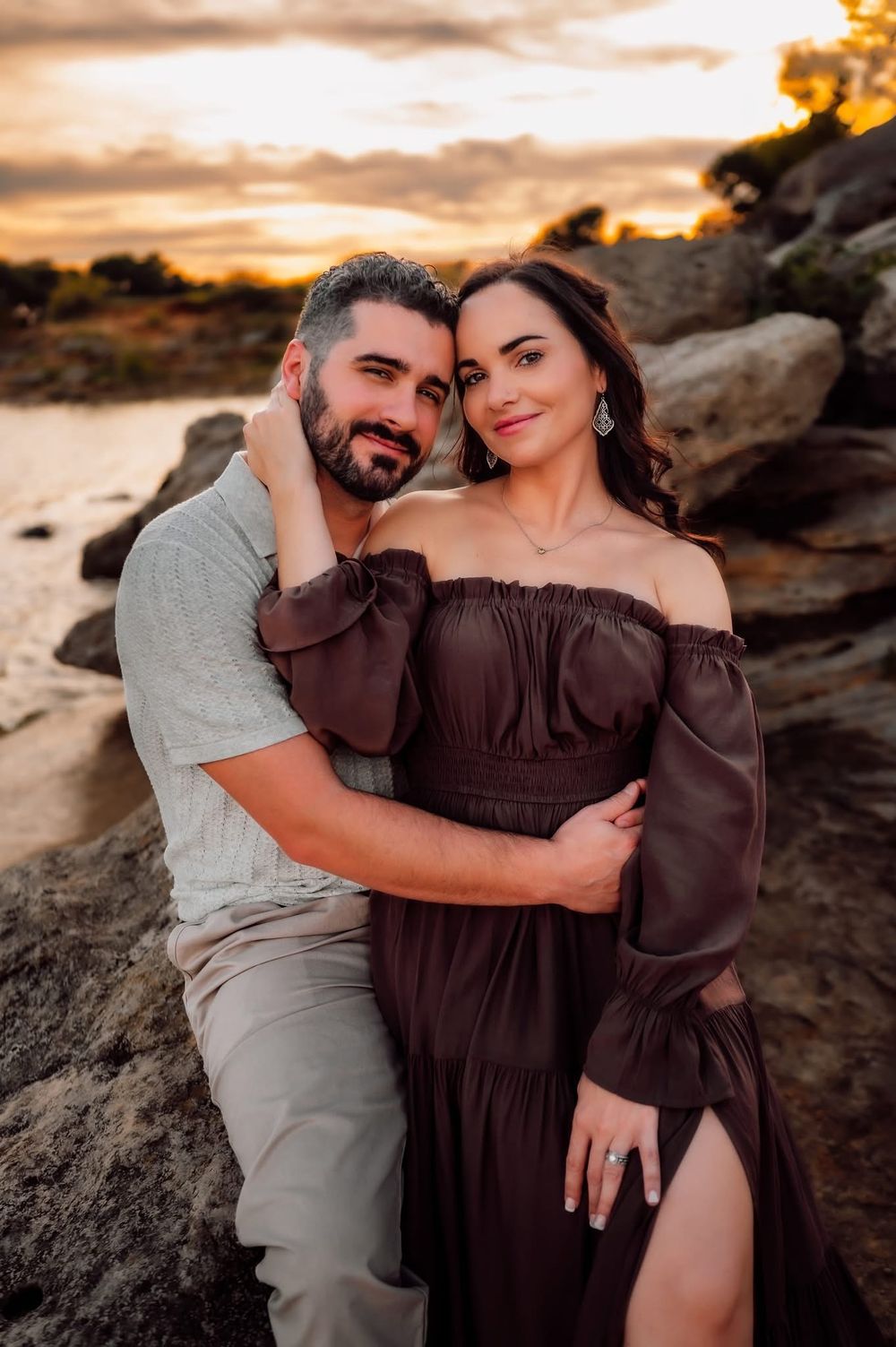 Couple embracing at sunset near rocky shoreline, woman in elegant brown off-shoulder gown with engagement ring.