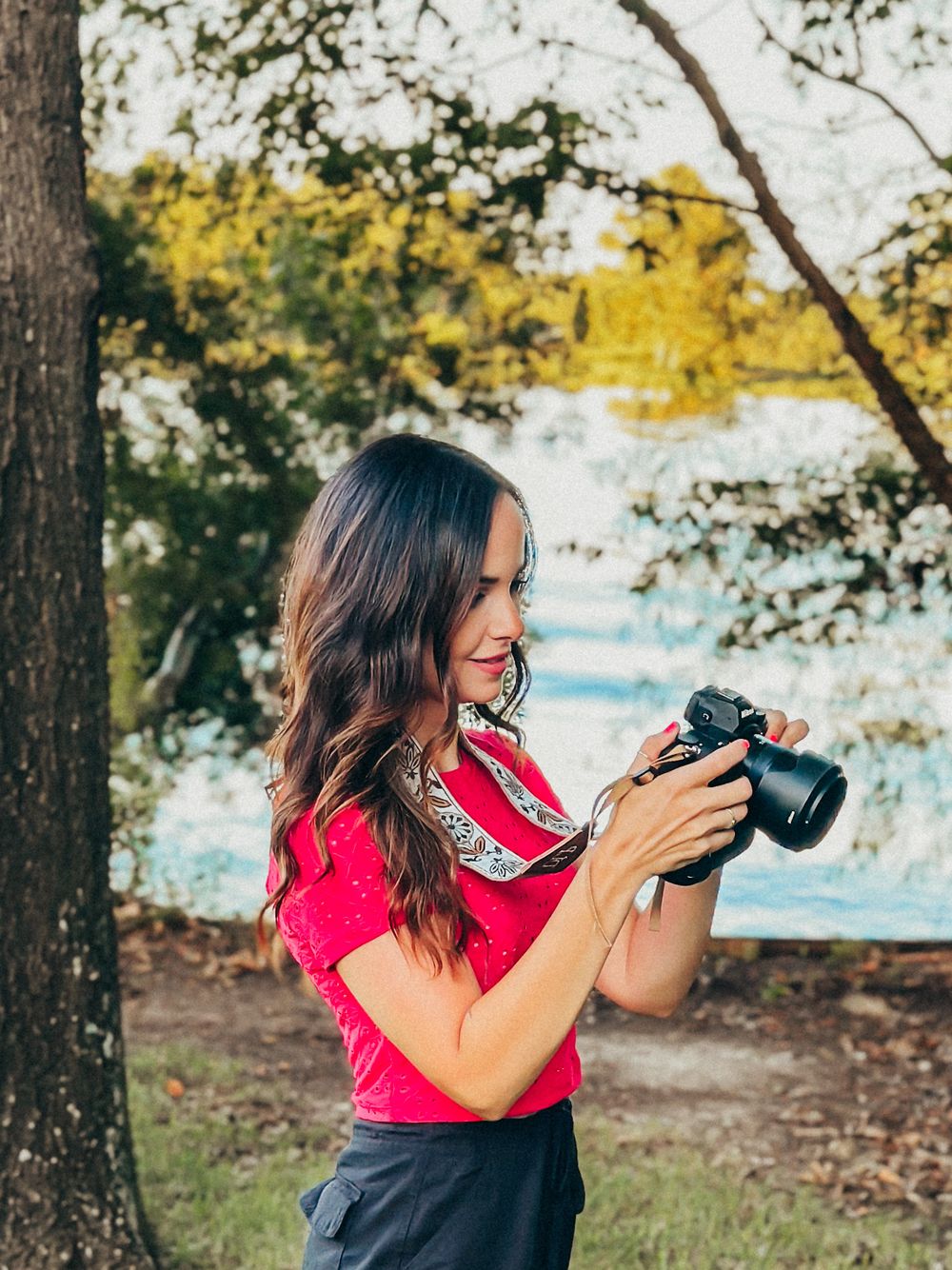 Female photographer holding DSLR camera outdoors near trees and water wearing a bright pink top.