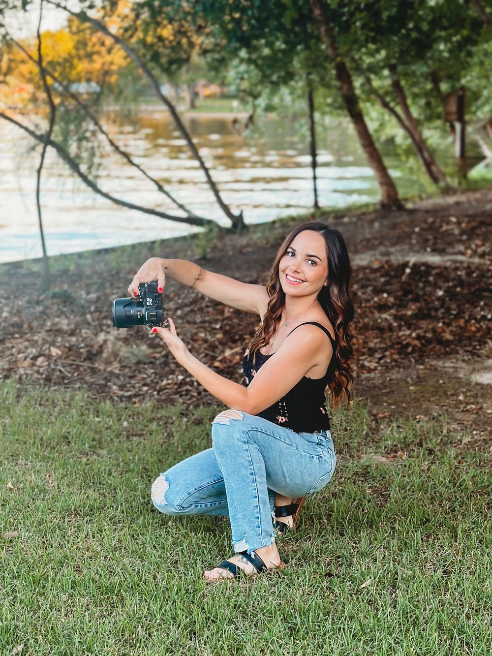 Female photographer crouching on grass holding camera near lakeside trees in jeans and black top.