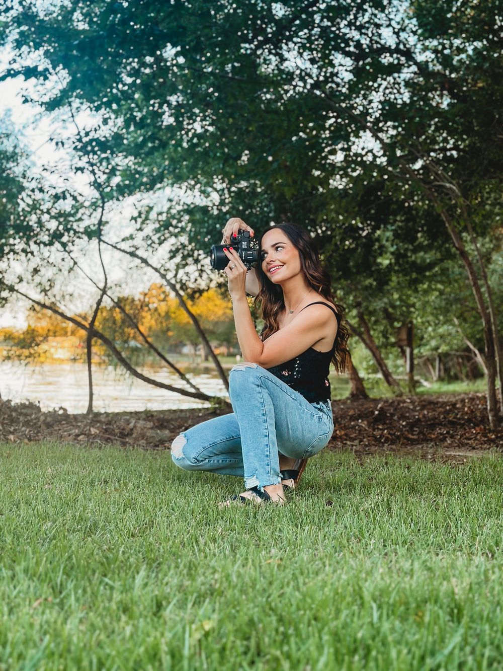 Female photographer crouching on green grass aiming DSLR camera near scenic lakeside tree backdrop.