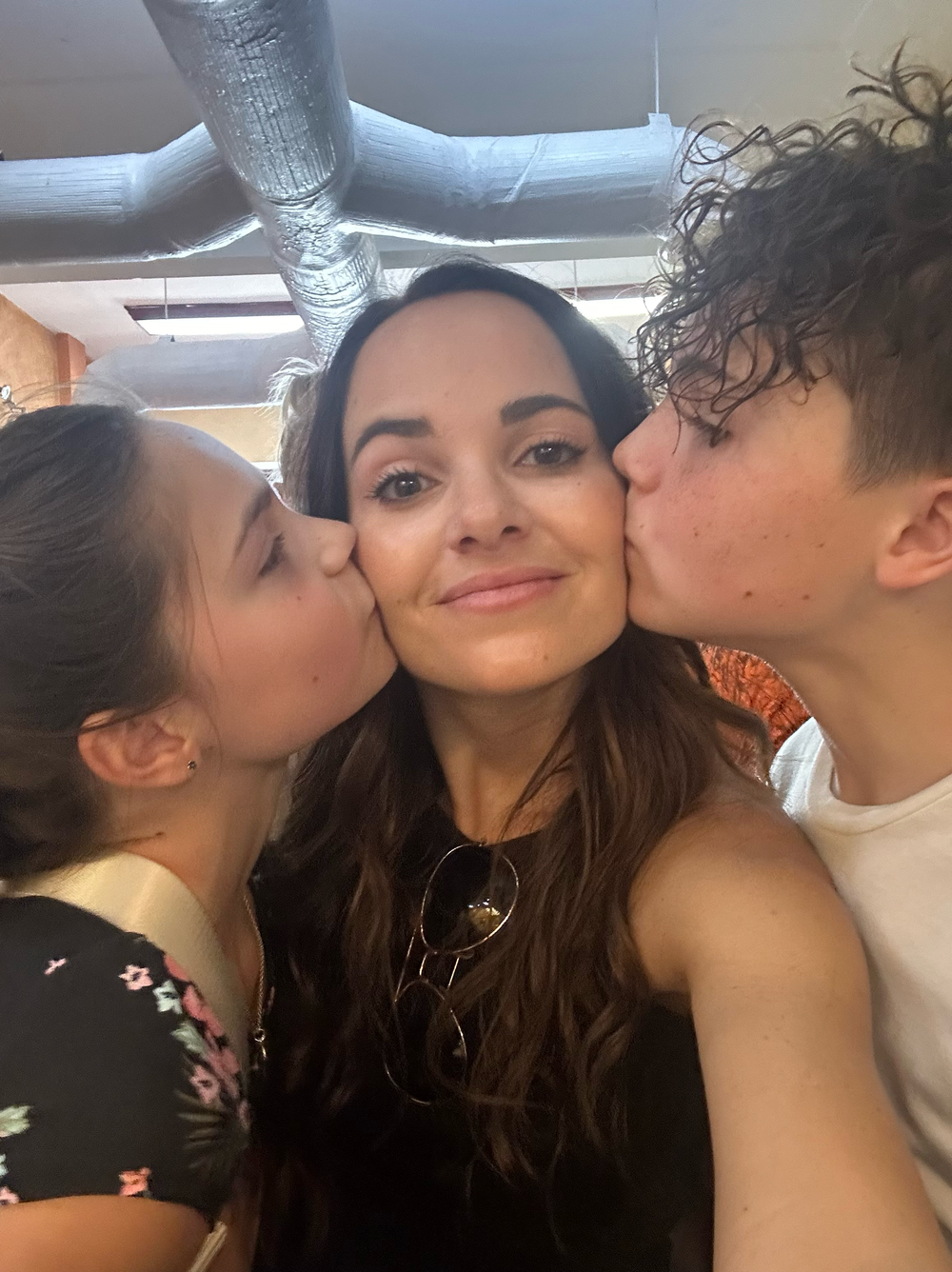 Woman smiling as two children kiss her on both cheeks in a sweet candid selfie moment.