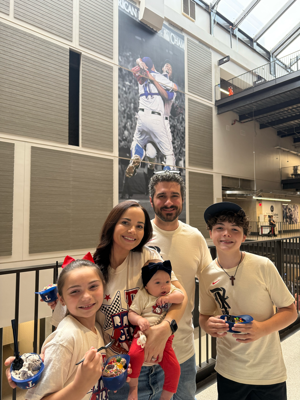 Family of five posing indoors at baseball stadium beneath large player mural with snacks and drinks.