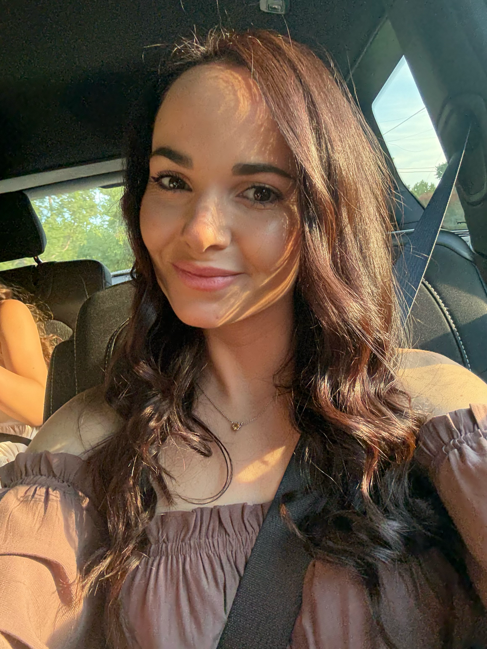 Young woman with long dark wavy hair taking a selfie in a car wearing a brown off-shoulder top.