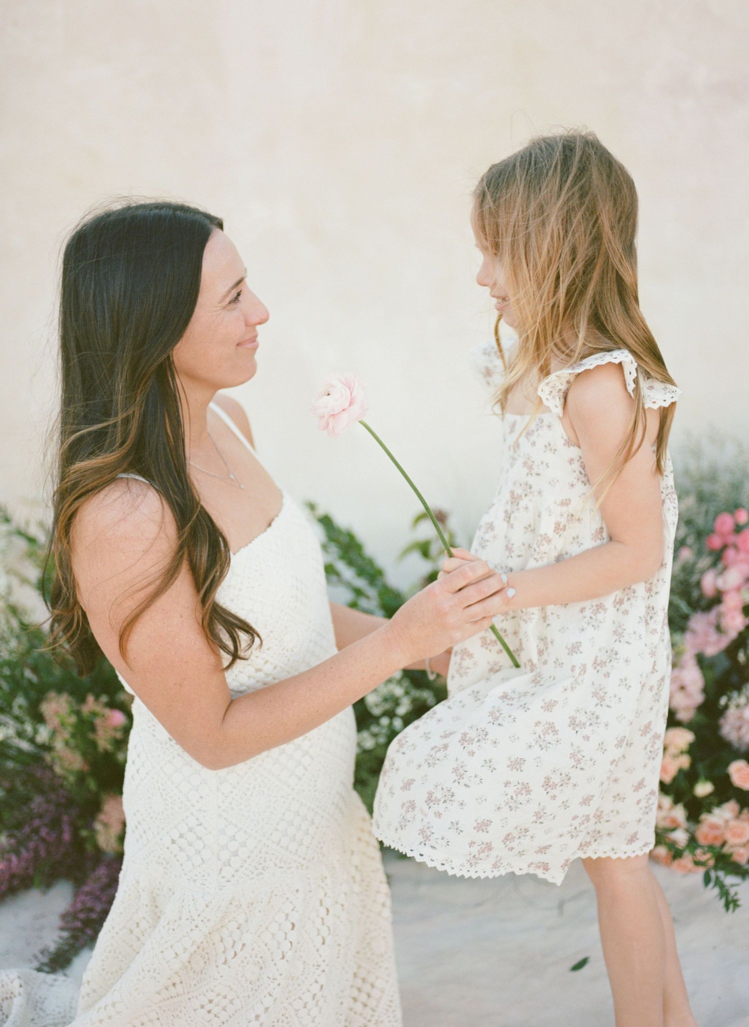 A mother in a white lace dress shares a pink flower with her young daughter in a floral dress outdoors.