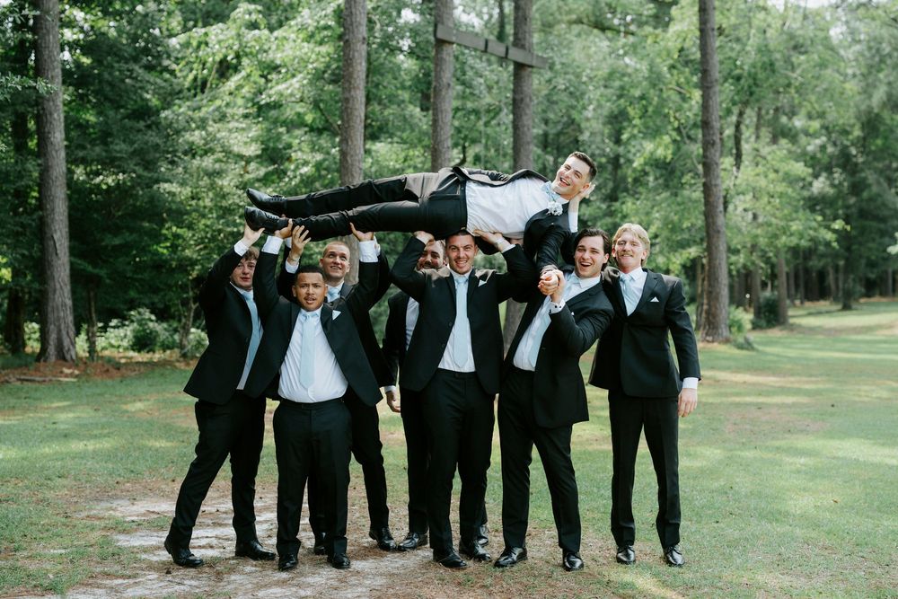 Groomsmen in black suits lifting the groom overhead outdoors in a wooded setting at a wedding celebration.