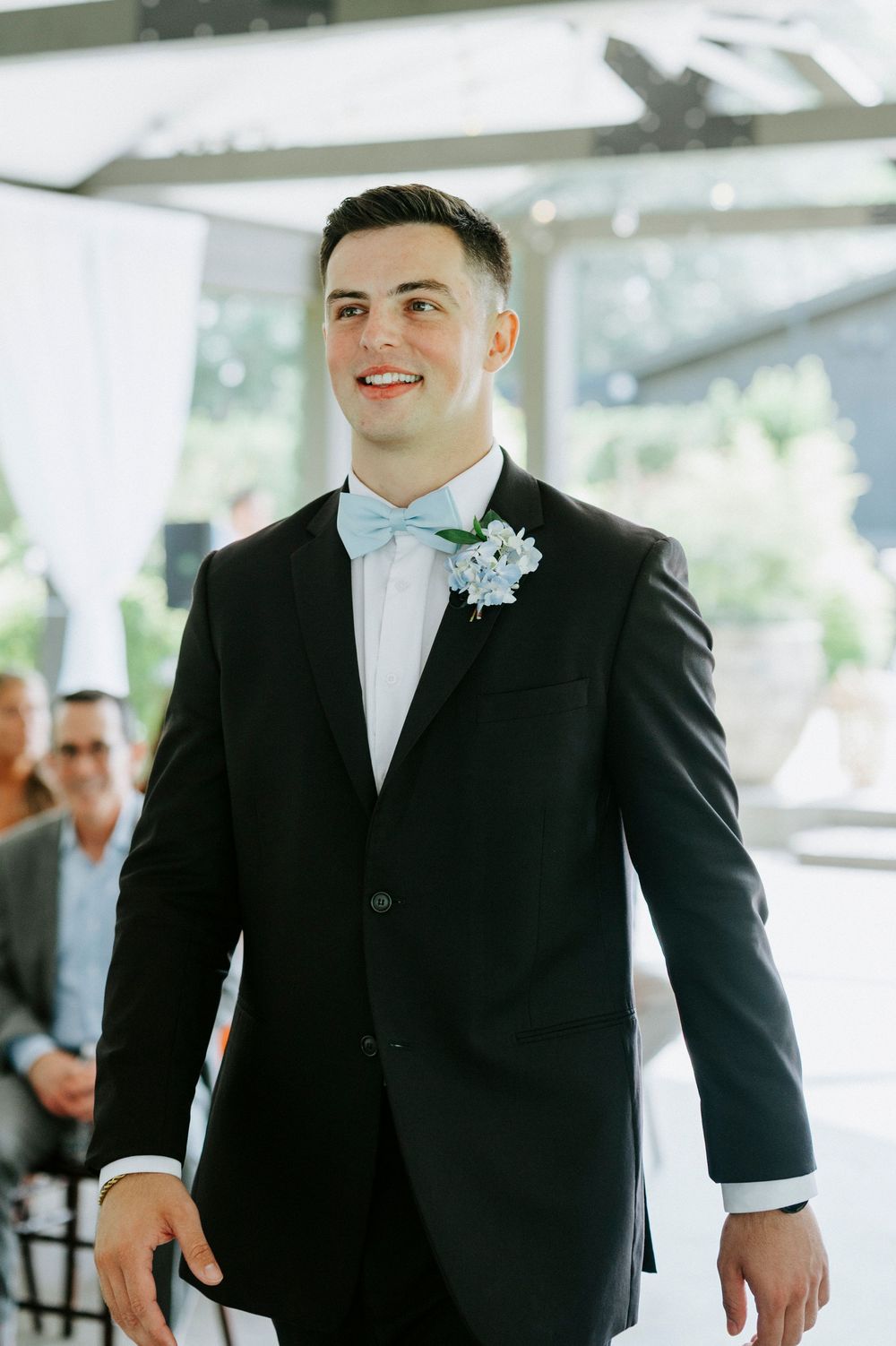 Smiling groom in black tuxedo with light blue bow tie and floral boutonniere walking at wedding ceremony.
