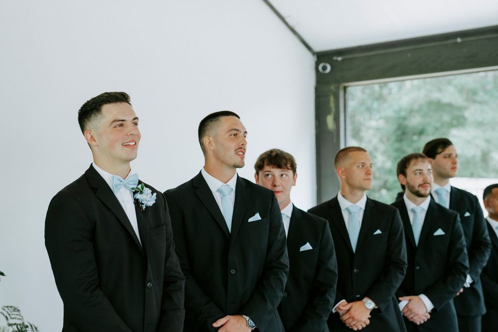 Groomsmen in black suits with white pocket squares smiling during a wedding ceremony.