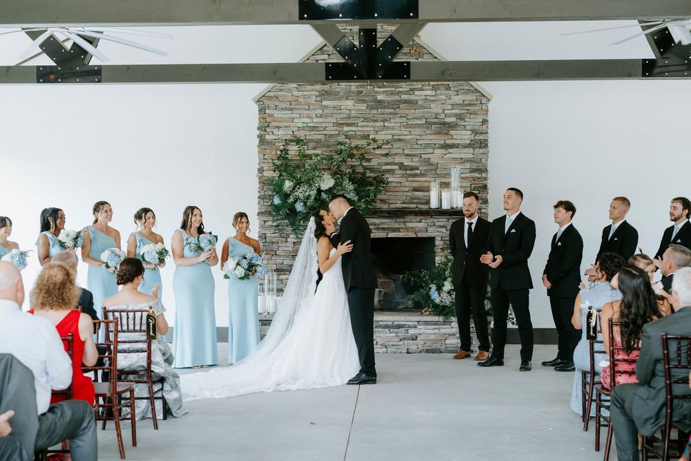 Bride and groom share first kiss at indoor wedding ceremony with stone fireplace and light blue bridesmaids.