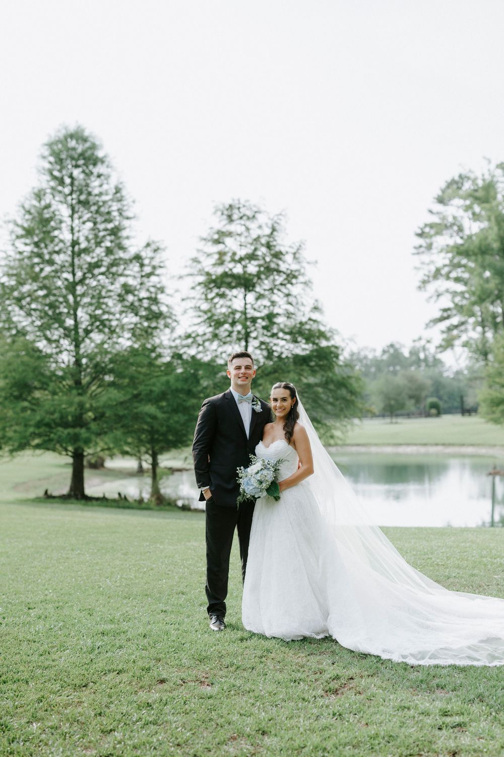 Bride and groom posing together on green lawn near a pond, surrounded by tall trees on their wedding day.