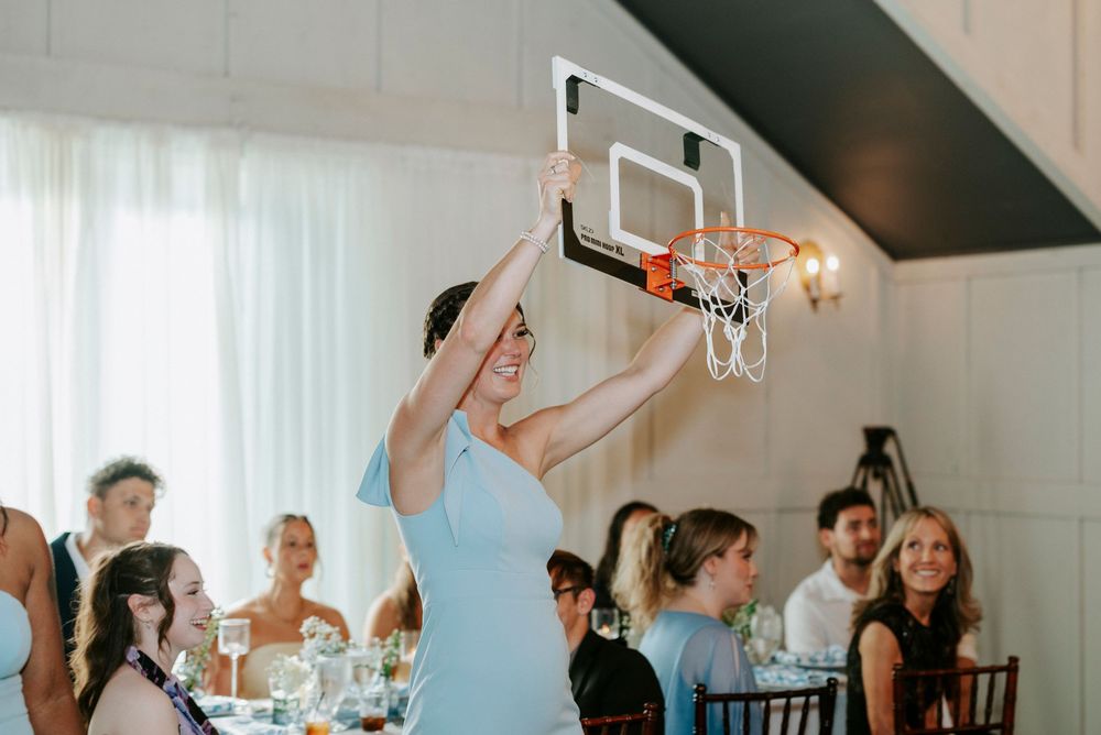 Woman in blue dress dunking basketball at indoor wedding reception with guests seated at tables.