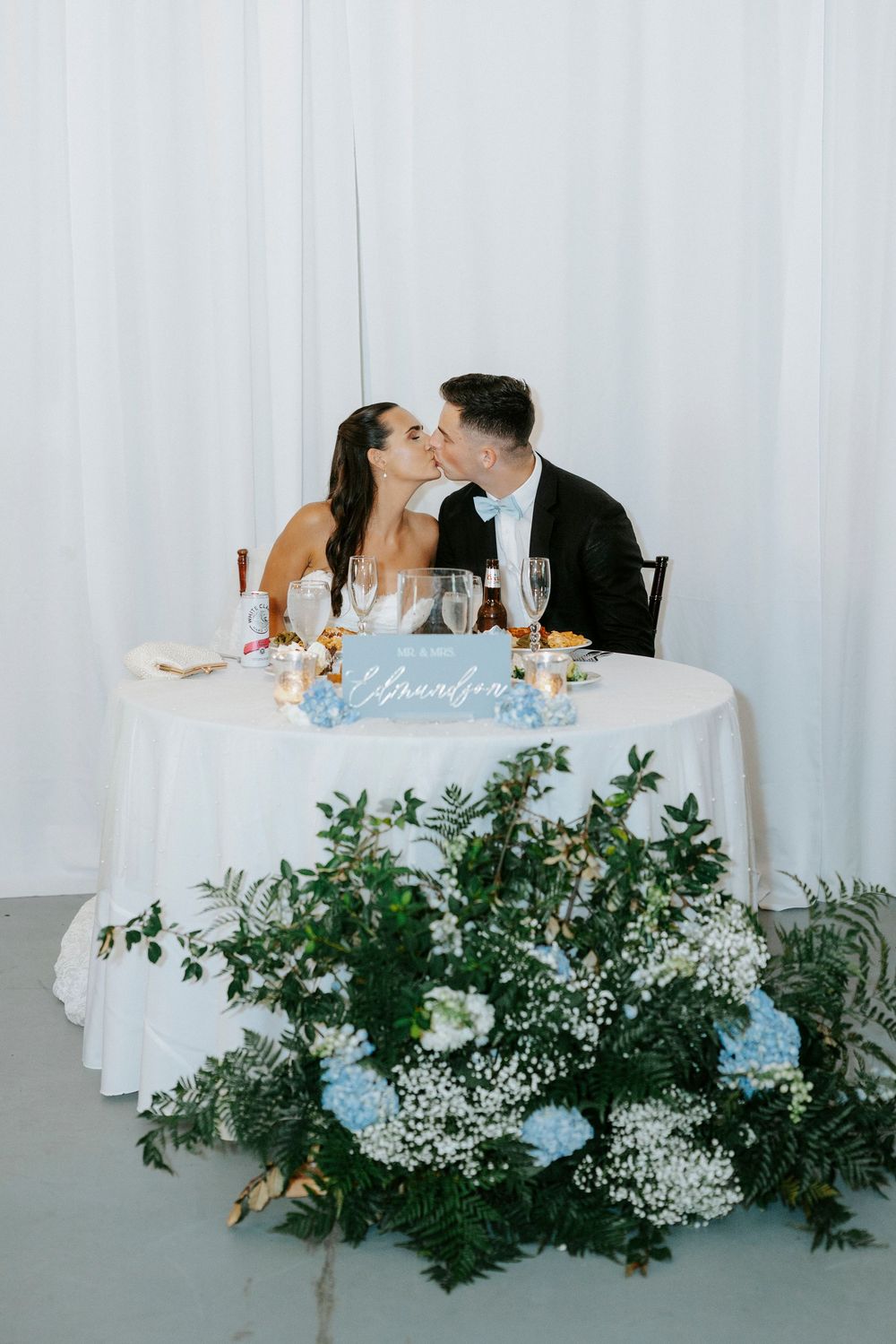 Newlyweds share a kiss at their sweetheart table adorned with blue hydrangeas, baby's breath, and lush greenery.