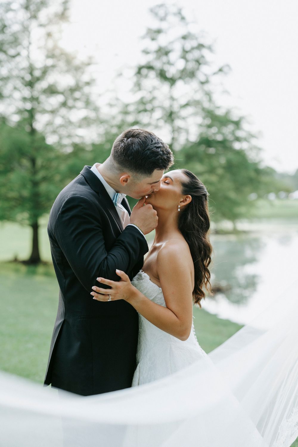 Bride and groom share a romantic kiss outdoors near a pond, with her veil flowing in the breeze.