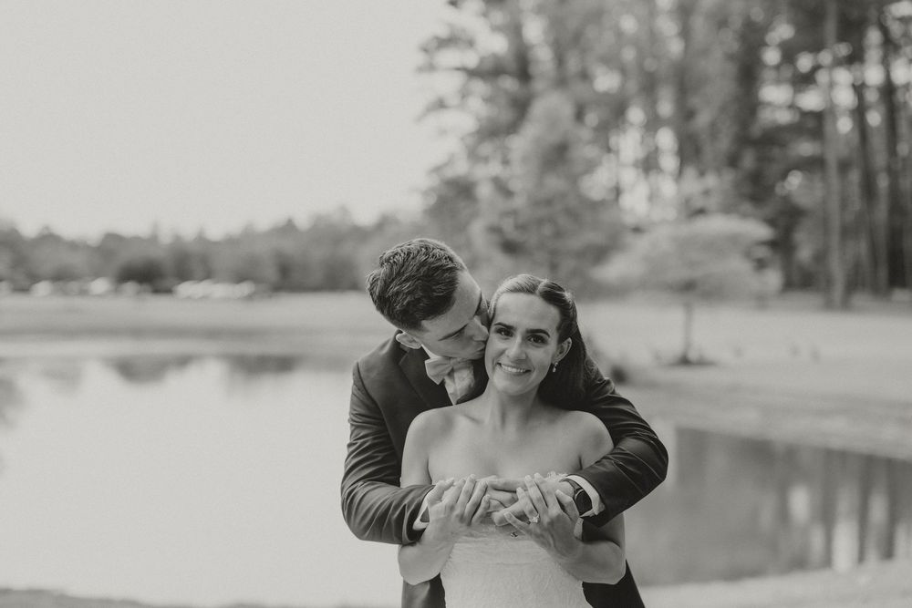 Black and white photo of a groom embracing his bride near a serene lake surrounded by trees.