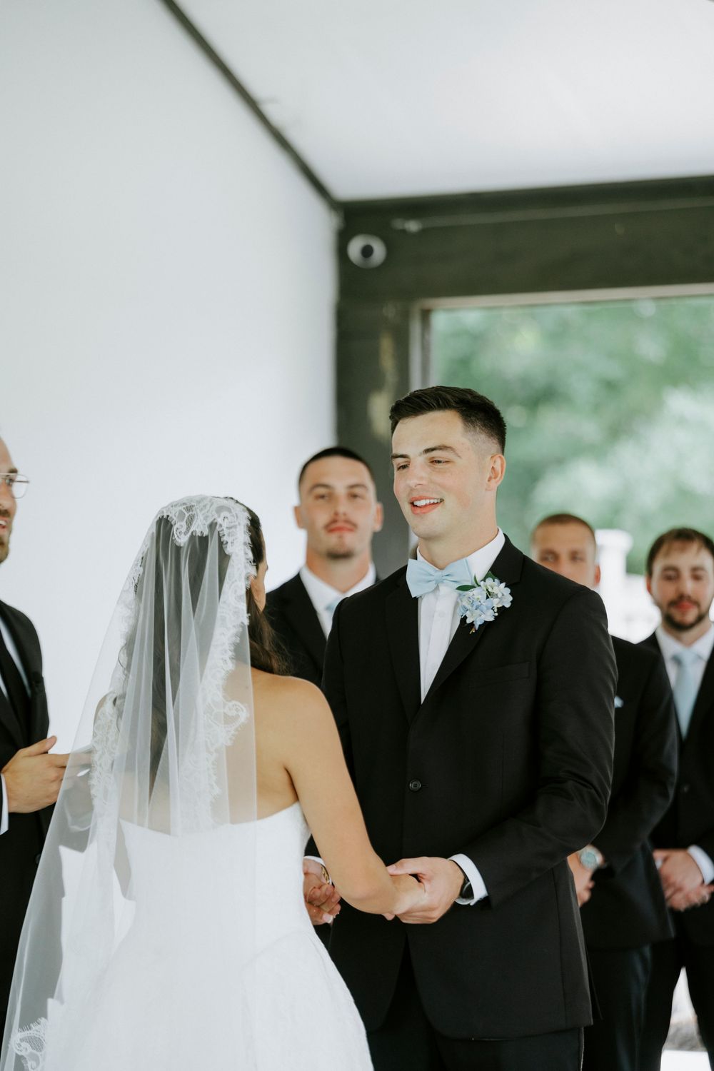 Groom in black tuxedo with blue bow tie smiles at bride in white wedding dress and veil during ceremony.