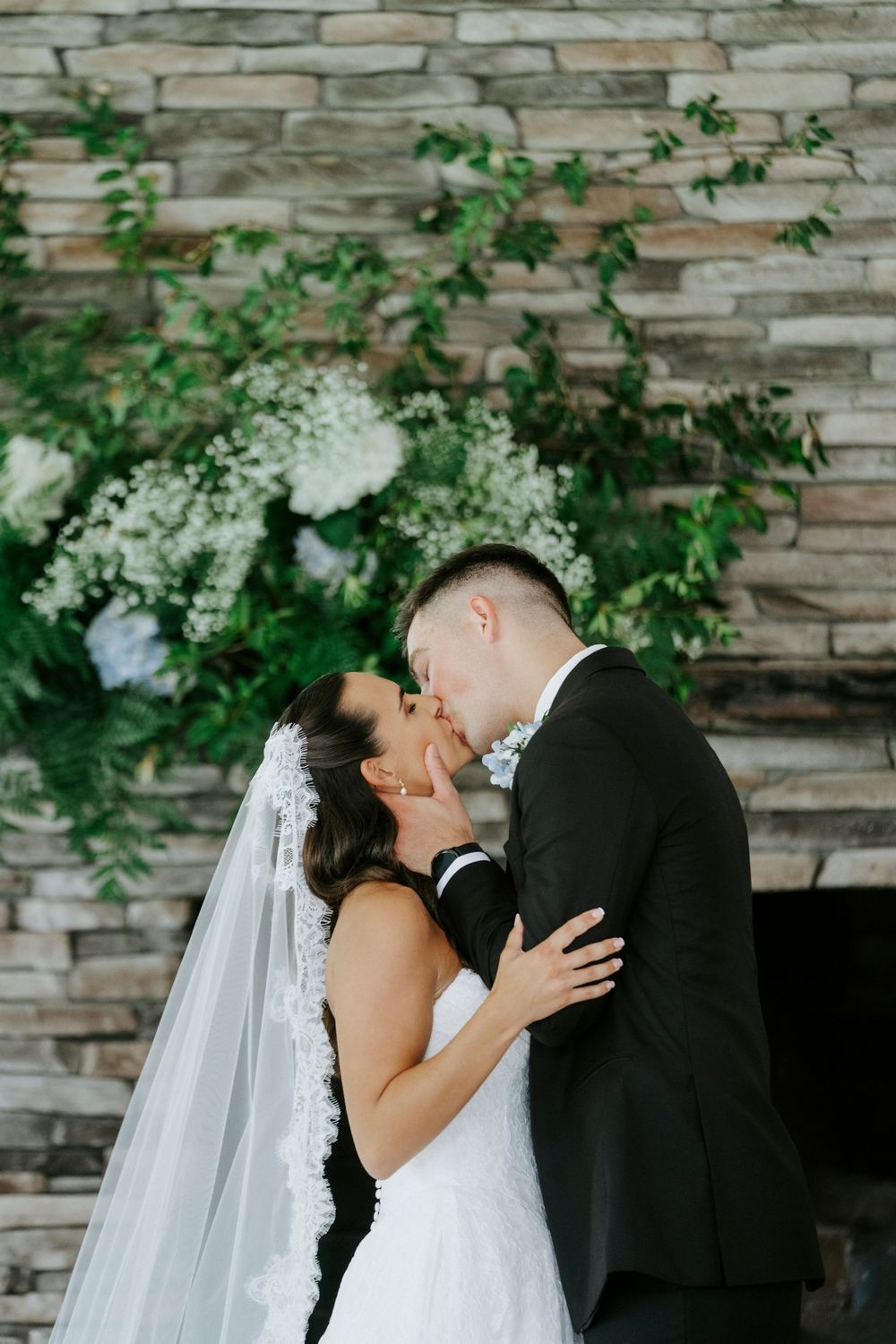 A bride and groom share their first kiss in front of a stone fireplace adorned with lush greenery and white flowers.