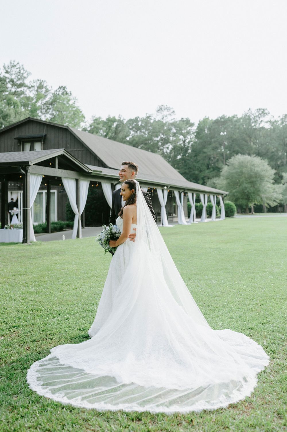 Bride in elegant white gown with long veil stands with groom on green lawn before a dark barn wedding venue.