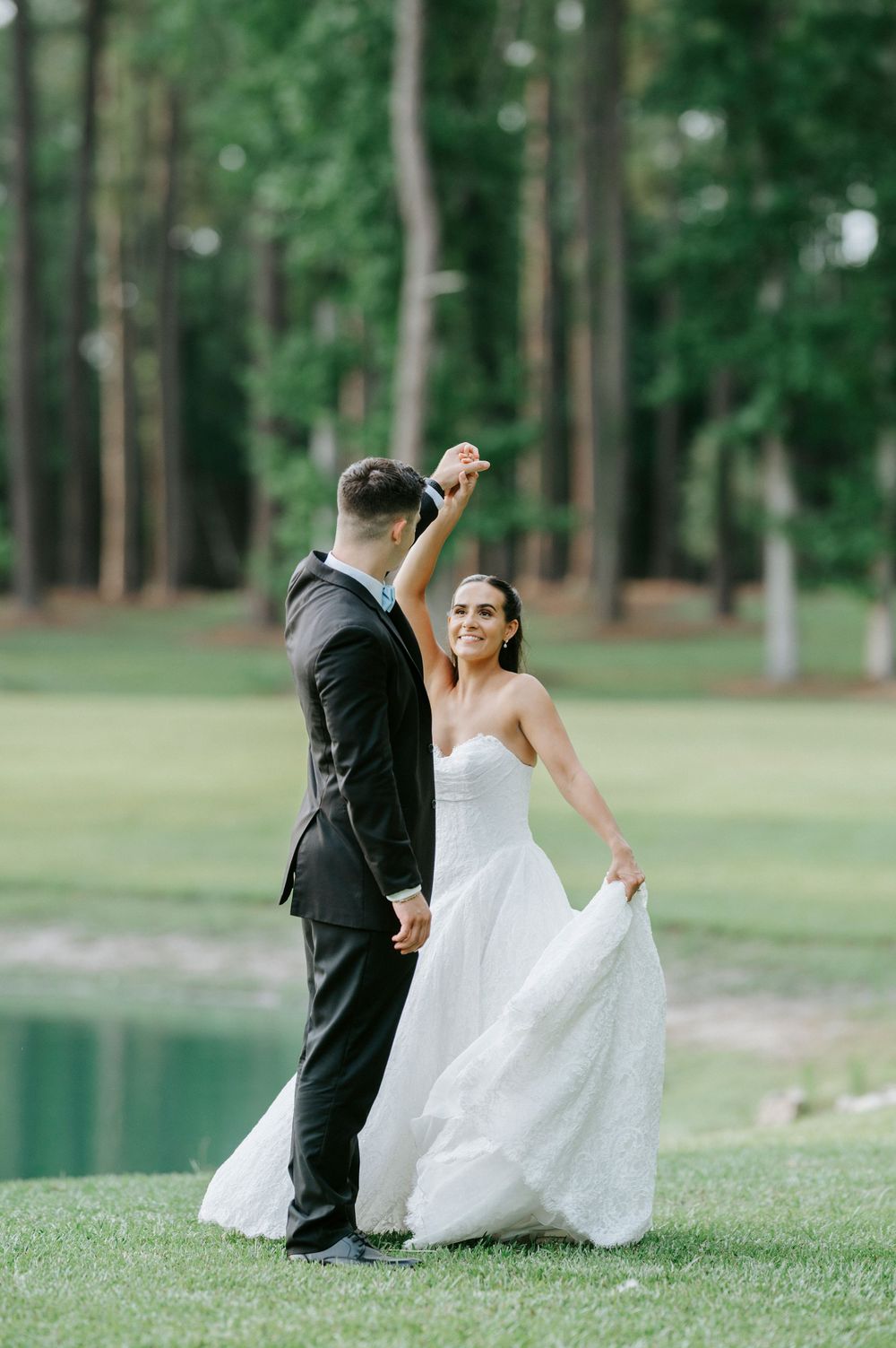 Bride and groom dancing outdoors near a pond, surrounded by tall trees, as she twirls in her white strapless gown.