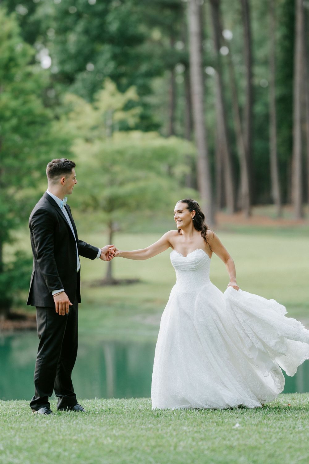 A bride in a flowing white gown dances with her groom in a black tuxedo on a lush green lawn near a pond.