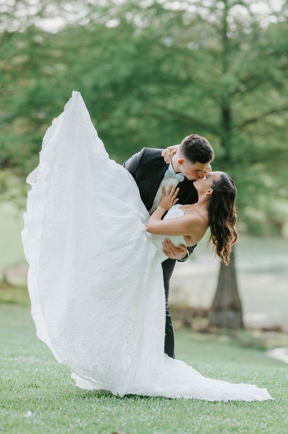 Groom lifts bride in white lace gown for a romantic dip kiss on green lawn with trees in background.