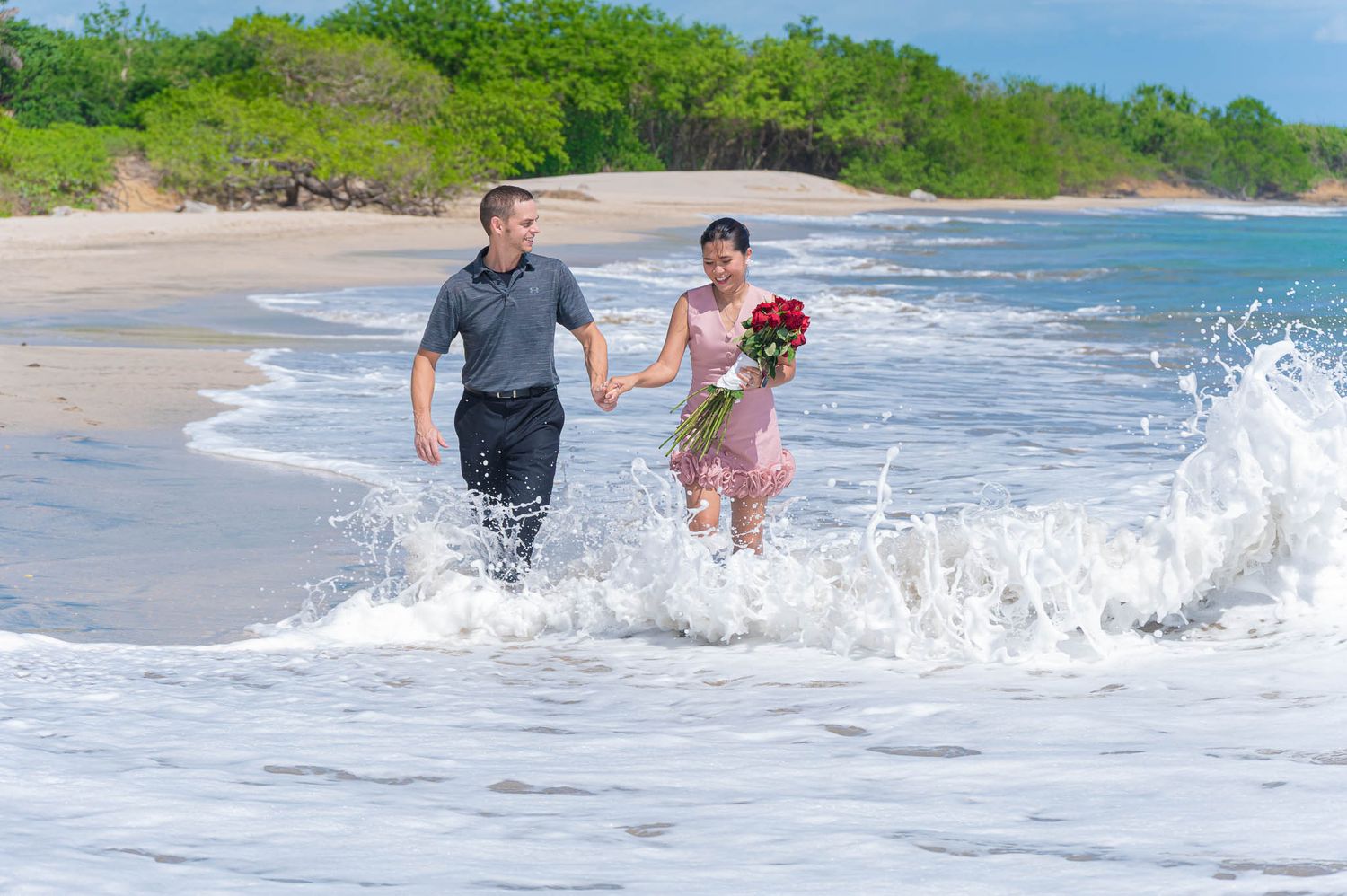 Couple holding hands and running through ocean waves on a tropical beach, woman carrying red roses in Guanacaste, Costa Rica
