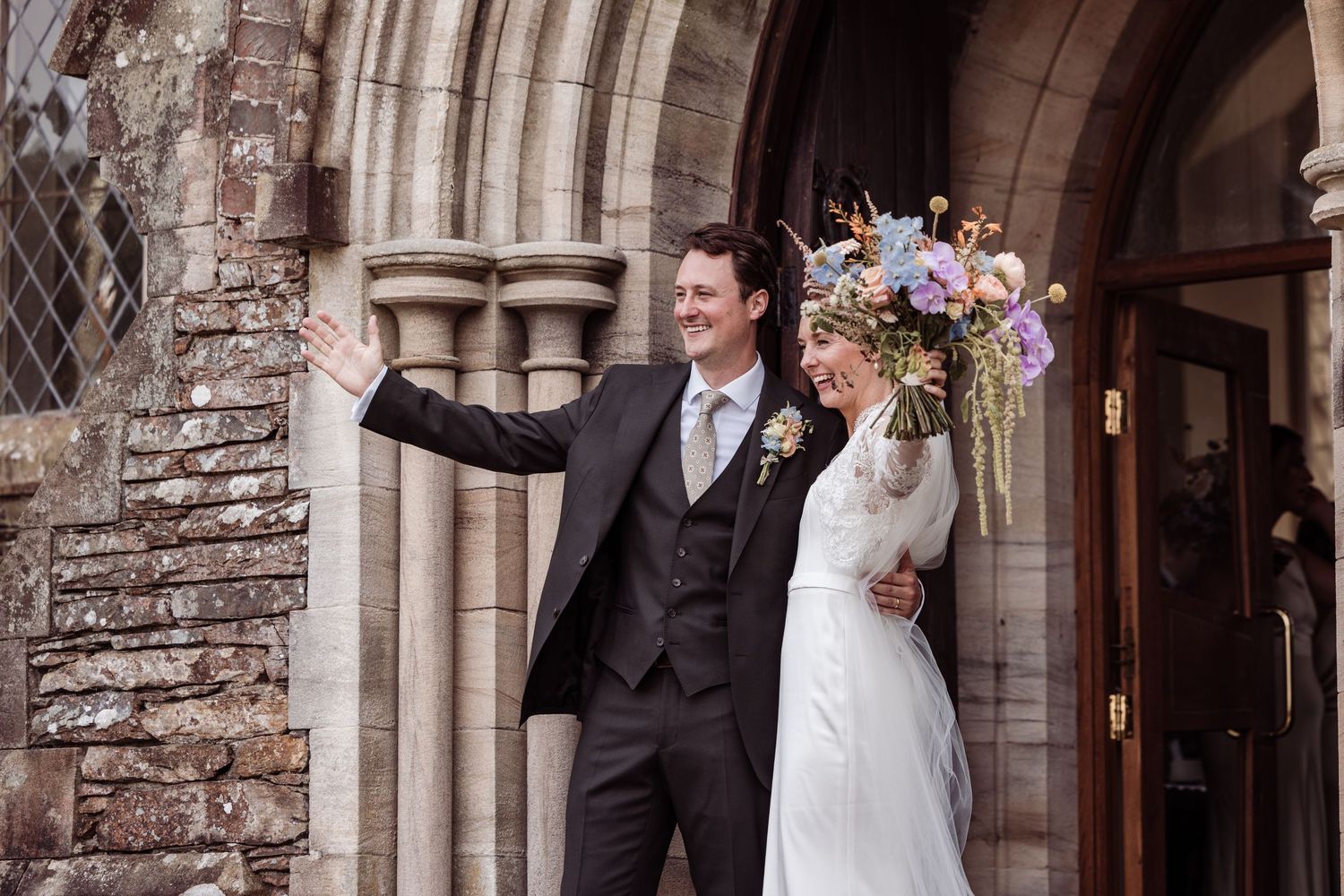 Newlywed couple celebrating outside a stone church, groom in dark suit waving, bride holding colorful wildflower bouquet.