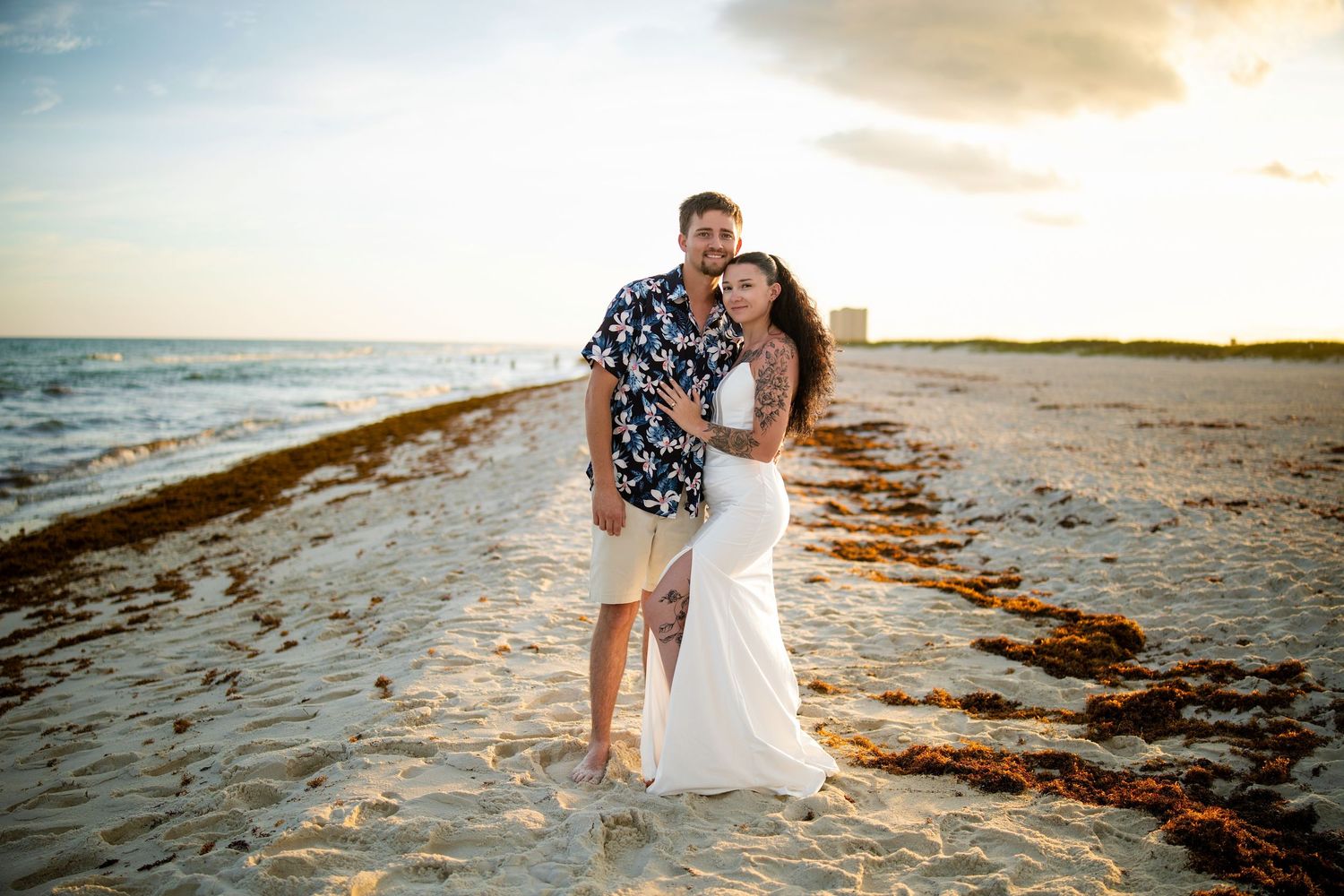 Couple embracing on a sandy beach at sunset, man in floral shirt and woman in white dress with ocean waves behind them.