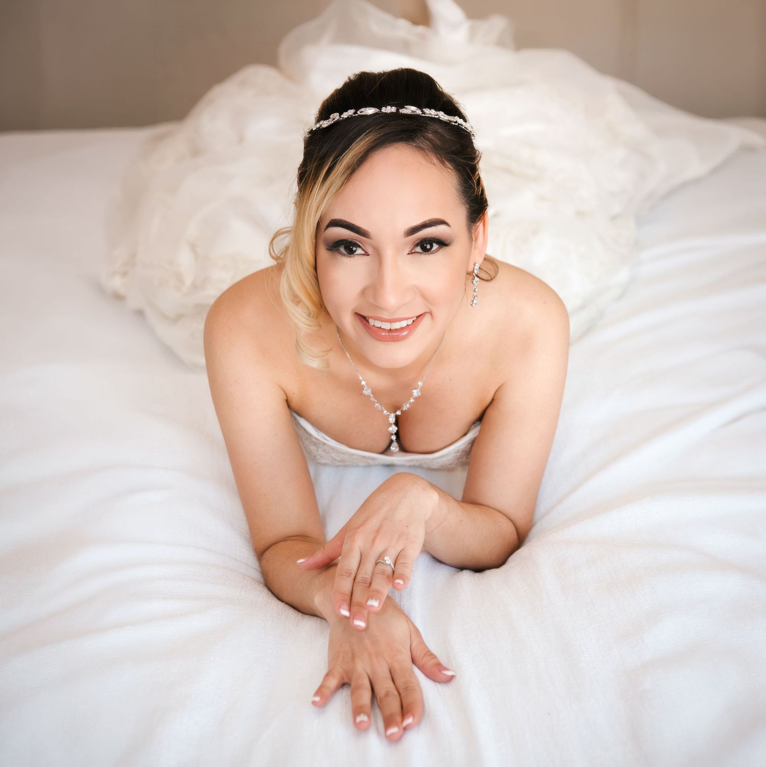 A smiling bride in a white gown and pearl headband poses gracefully on a white bed, wearing elegant jewelry.