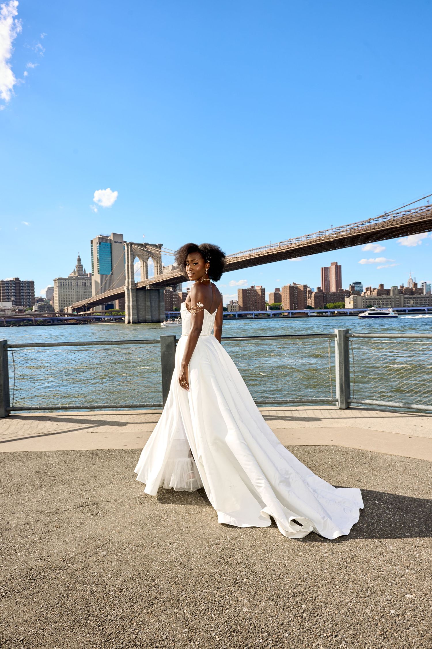 Black bride in white strapless ball gown wedding dress standing in lush green park, smiling downward.
