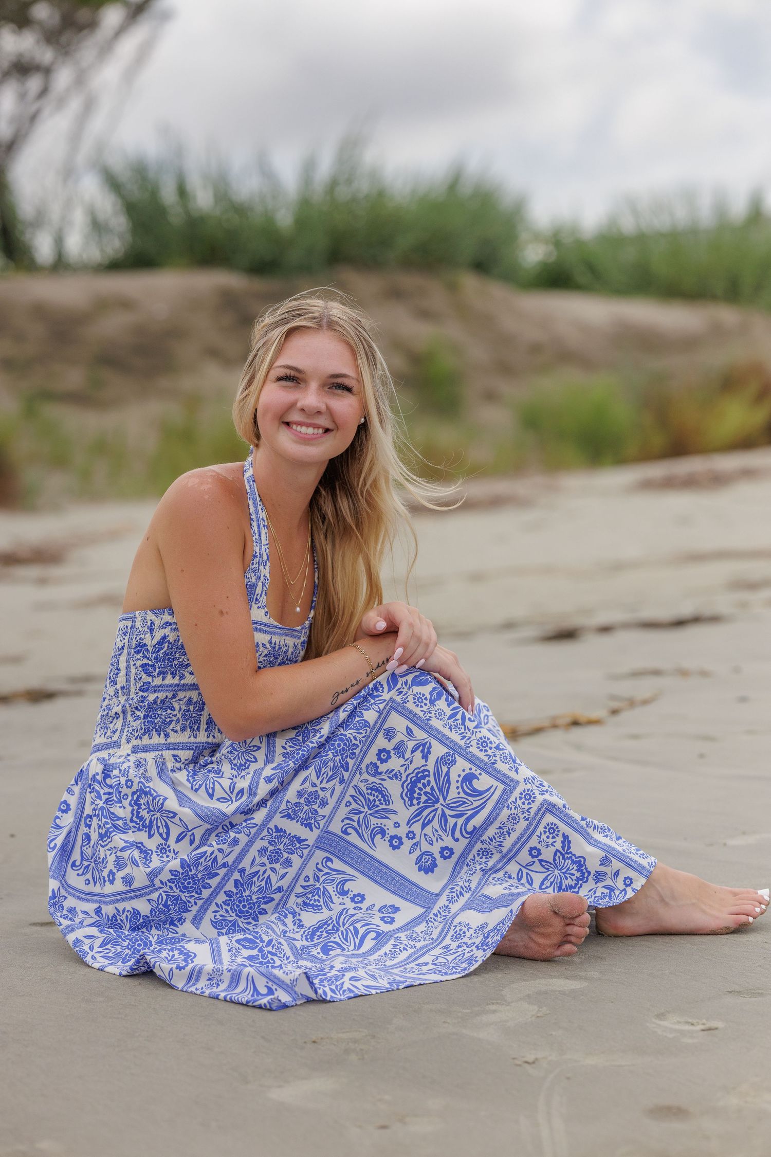Smiling blonde woman in blue floral sundress sitting barefoot on sandy beach with green dunes in background.