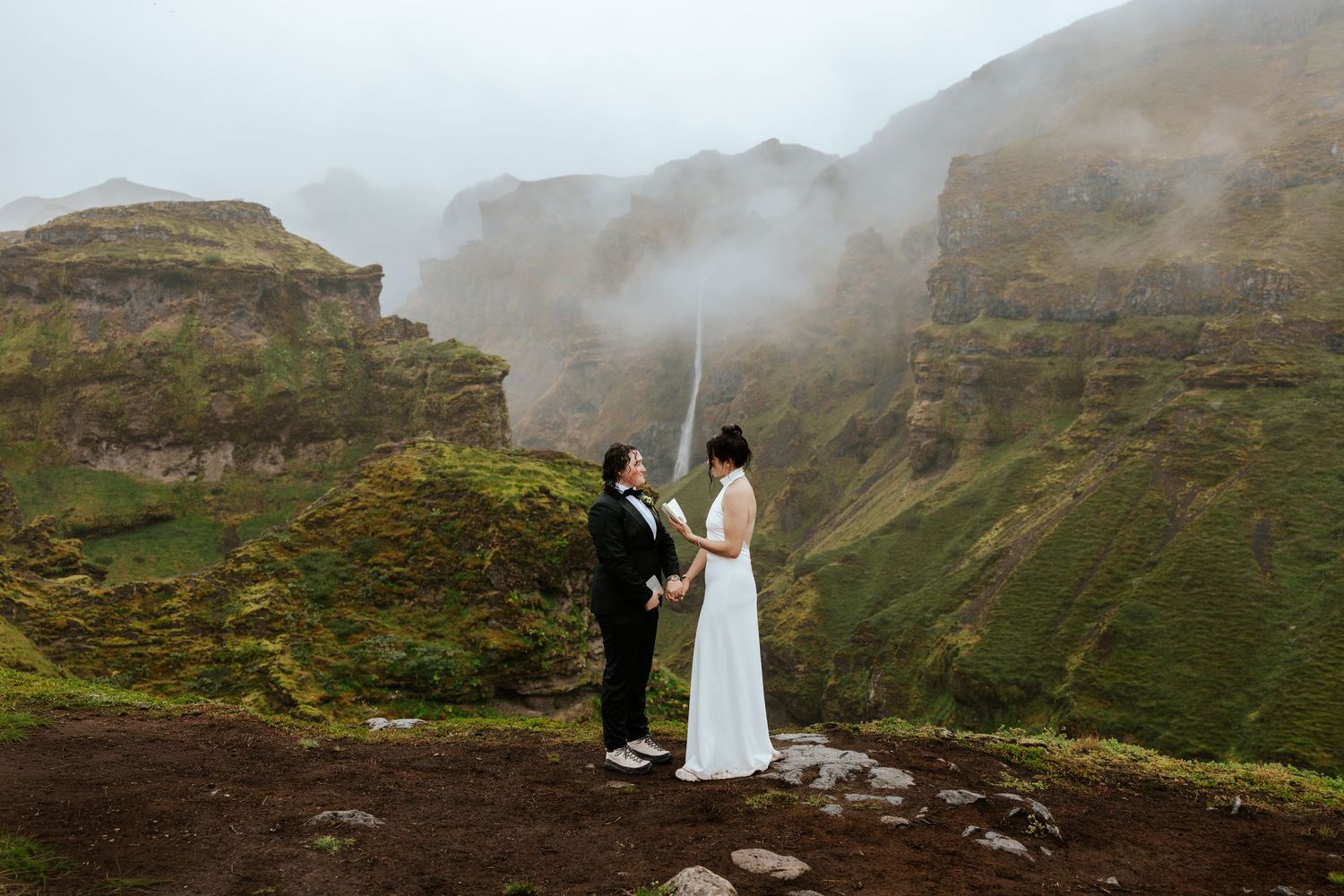 two brides standing on a mountain with a view of a canyon and waterfall while one bride reads vows at Mulagljufur Canyon, Iceland