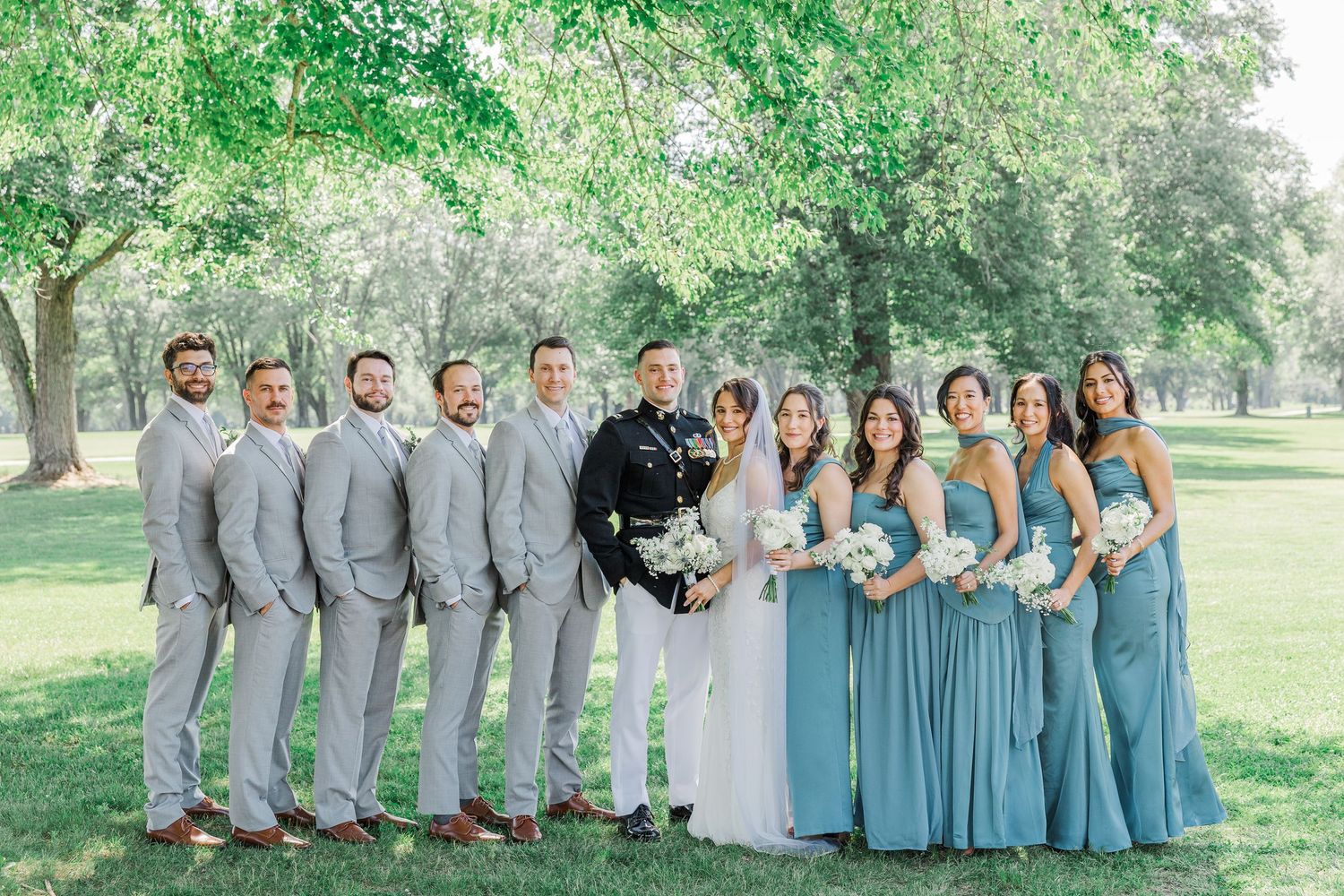 Wedding party poses outdoors under trees, groomsmen in gray suits and bridesmaids in dusty blue gowns flanking the bride and groom.