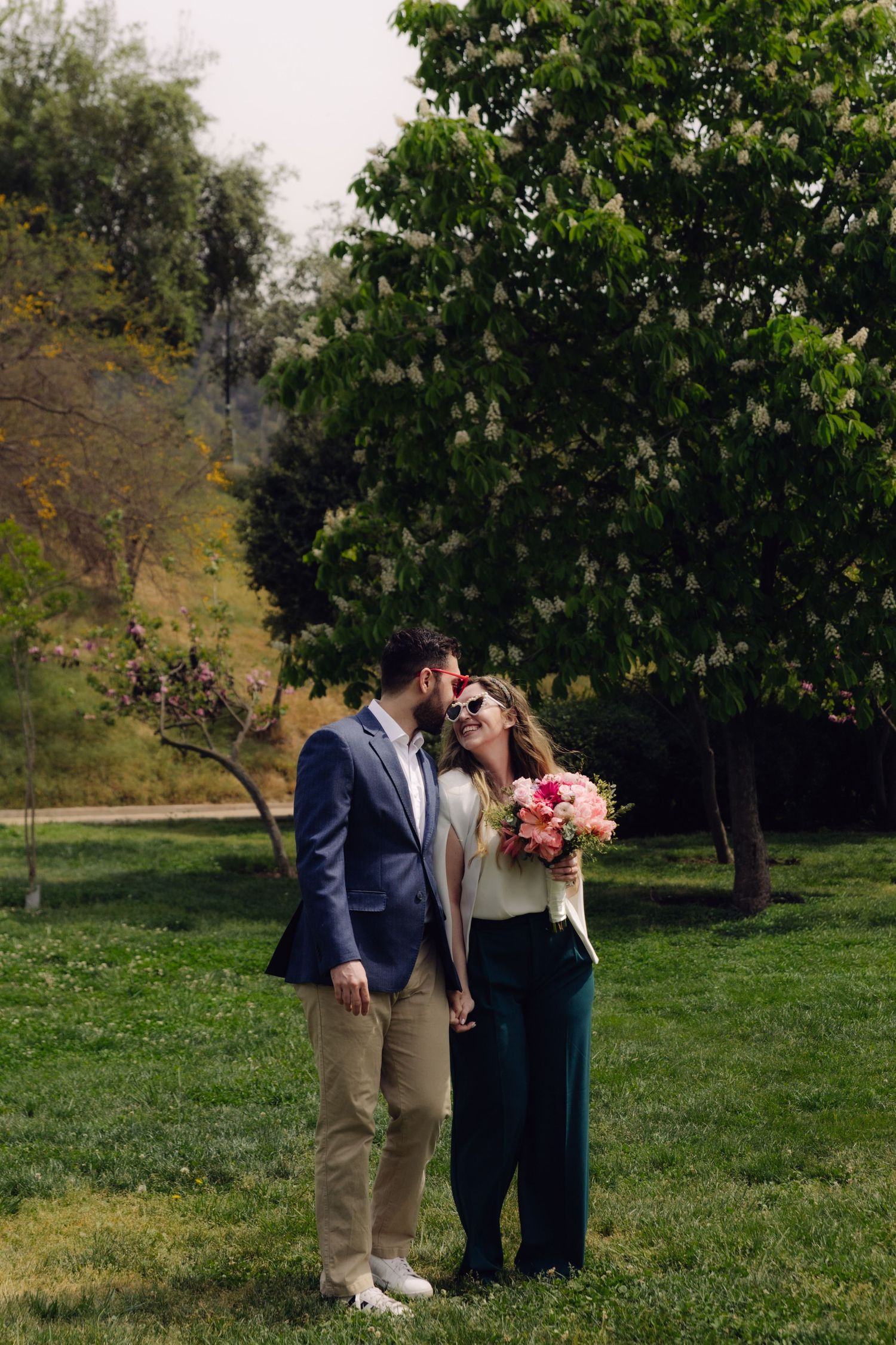 Bearded man in blue blazer kissing woman in white blazer holding pink bouquet in park.